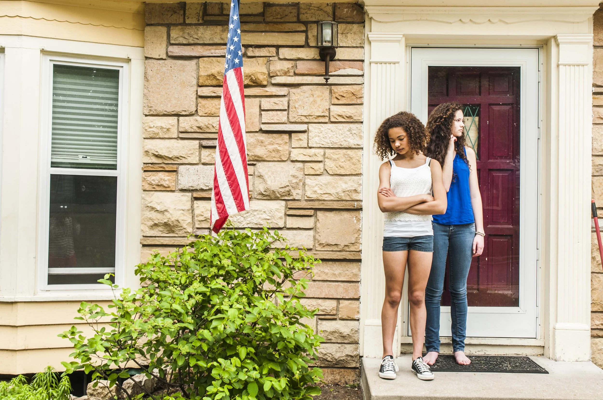 Two young women stand on a porch, looking down with somber expressions. One has short curly hair and is wearing a white tank top and shorts, the other has long curly hair and is wearing a blue sleeveless top and jeans. An American flag is displayed o