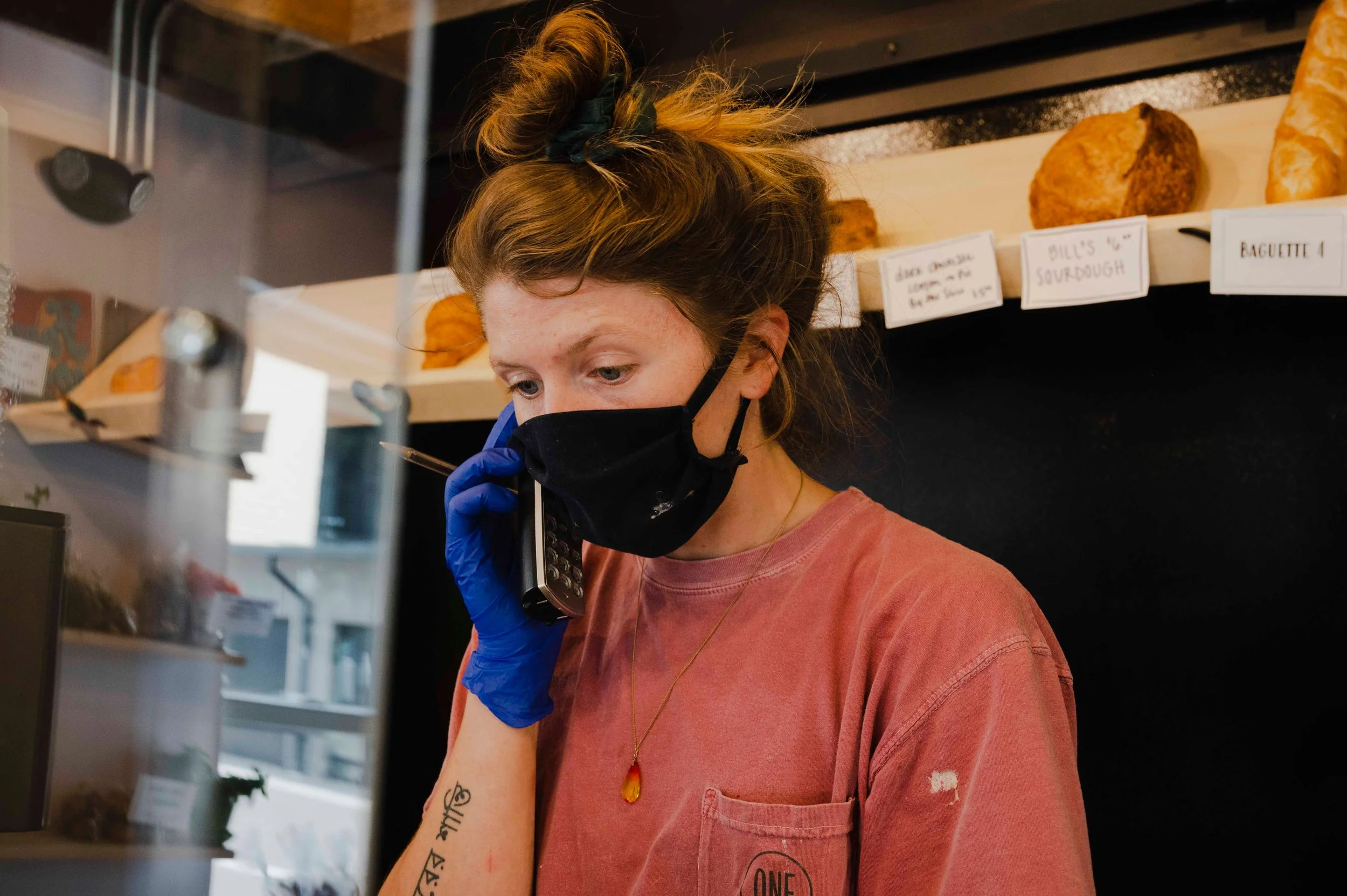 A woman with a red hair bun, wearing a black face mask and a blue glove, is talking on a landline phone inside a bakery or cafe. She is wearing a pink T-shirt and a gold necklace, with a tattoo on her left forearm, and there are bread loaves on a she