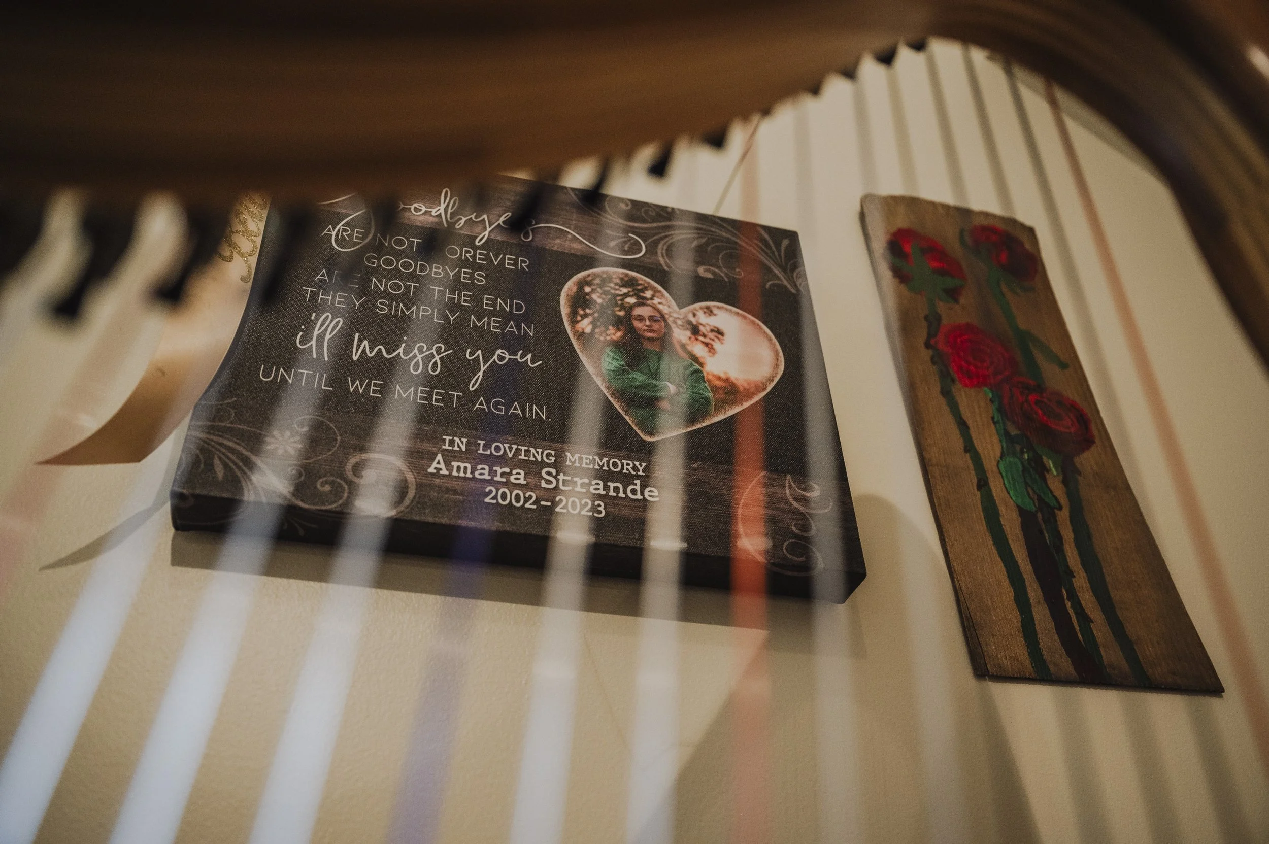 Memorial plaque with a photo of a young woman in a heart-shaped frame, with the words 'I miss you until we meet again,' and the name Amara Strande 2002-2023, surrounded by decorative swirls, against a dark background. Next to a wooden artwork of red 