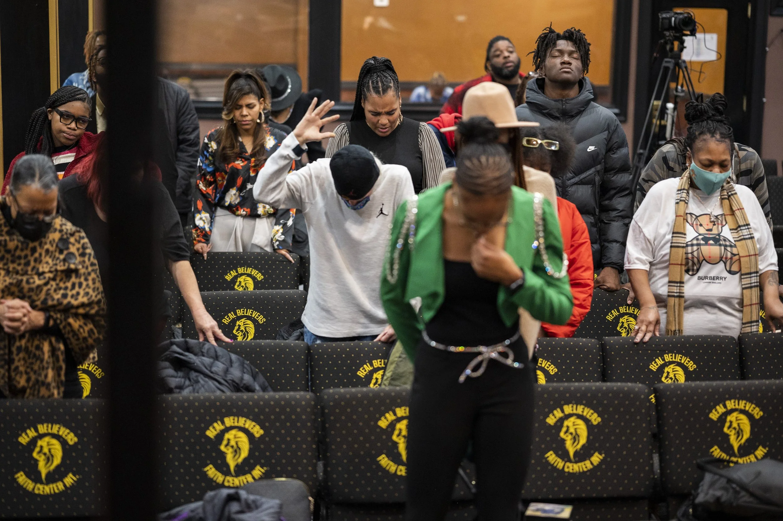A group of diverse people praying or meditating with eyes closed or bowed head, in a room with black chairs that have a yellow lion logo and text reading 'Real Believers Truth Center Inc.'