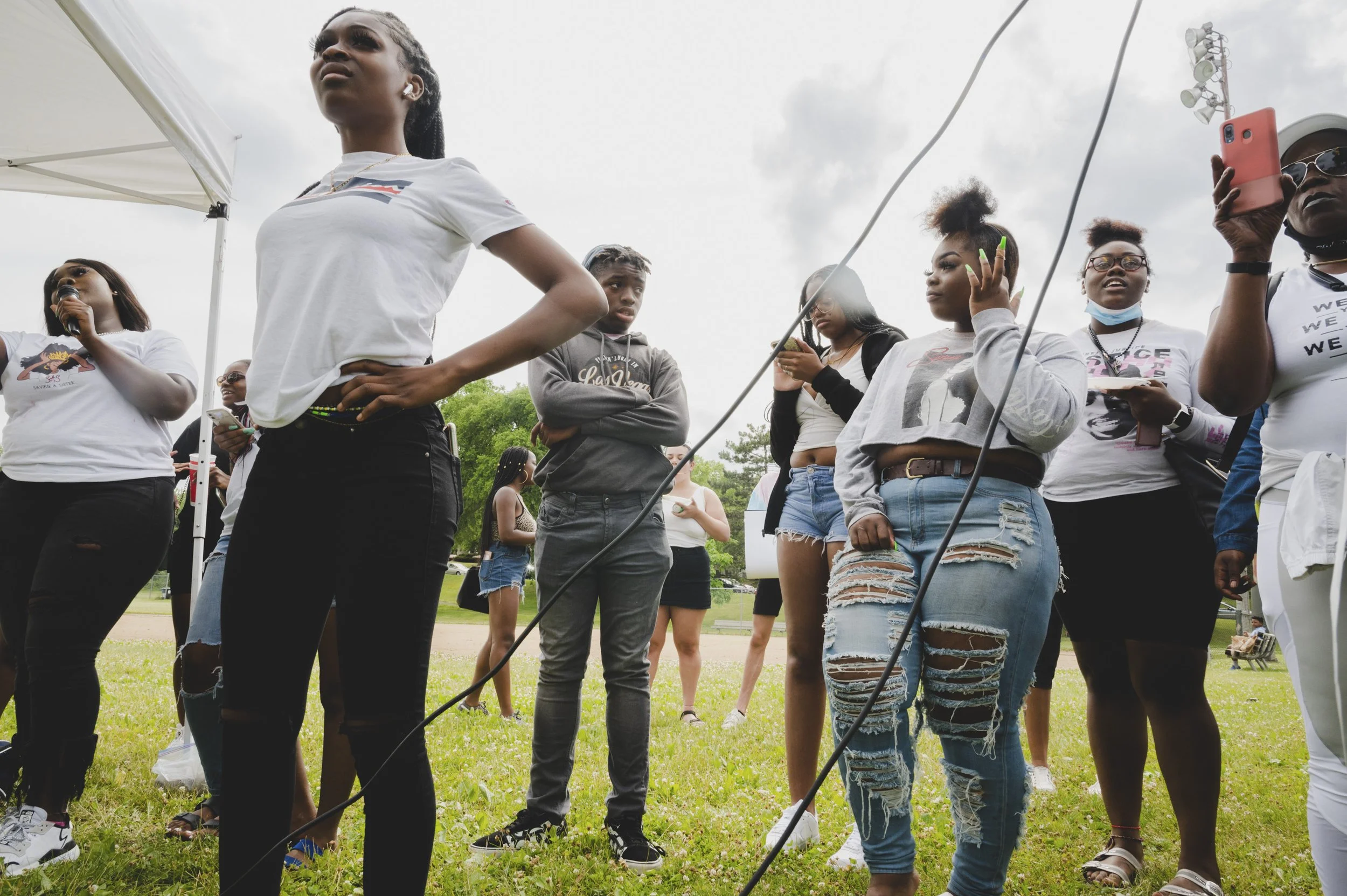 Group of young women standing outdoors on grass, some using their phones, some wearing white shirts and sunglasses, during a gathering or event in daytime with overcast sky.