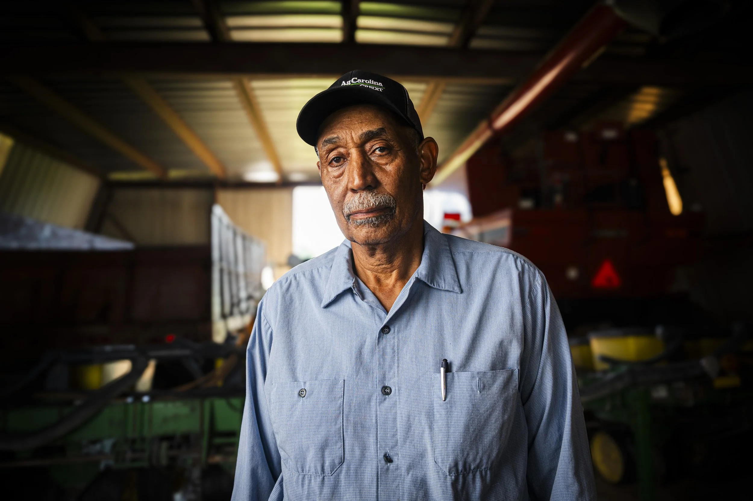 Portrait of a Black cotton farmer (an older man in a light blue button-up shirt) and black cap inside a barn or shed, with farm equipment and machinery in the background.