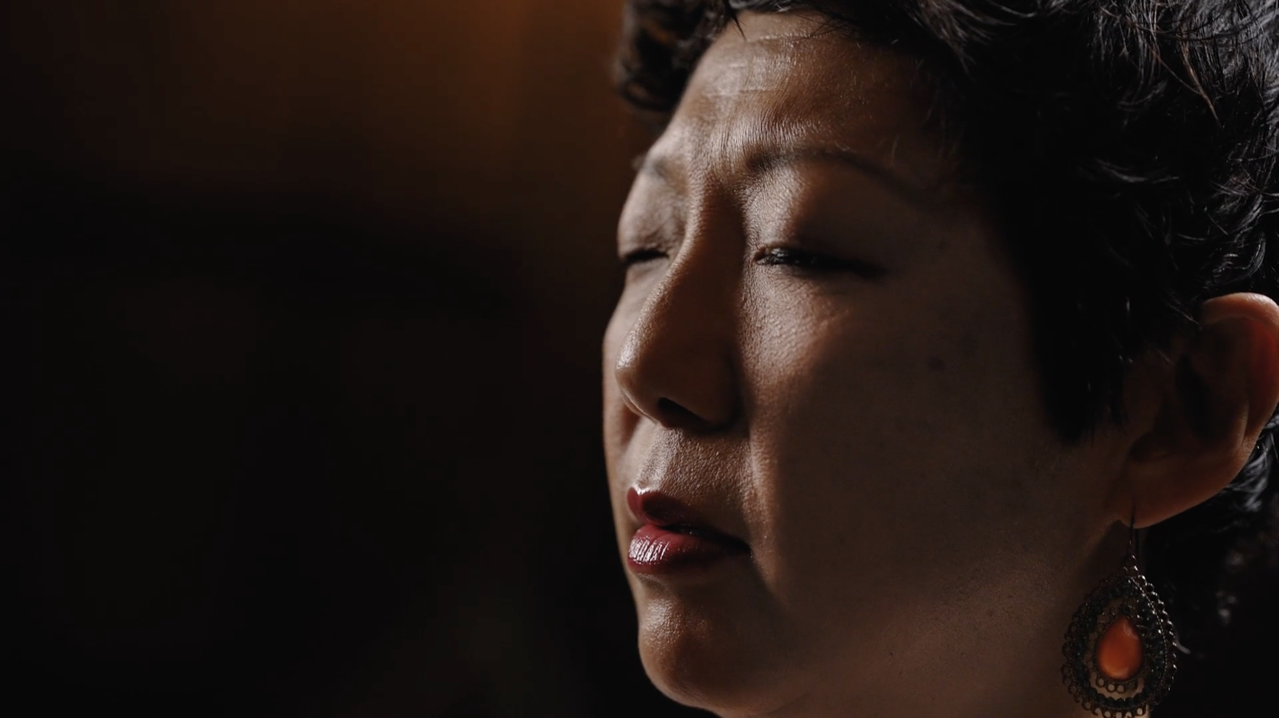 Close-up of an Asian woman with closed eyes, dark curly hair, wearing red lipstick and a large orange and black earring, with a dark background. She is practicing a breathing excercise.