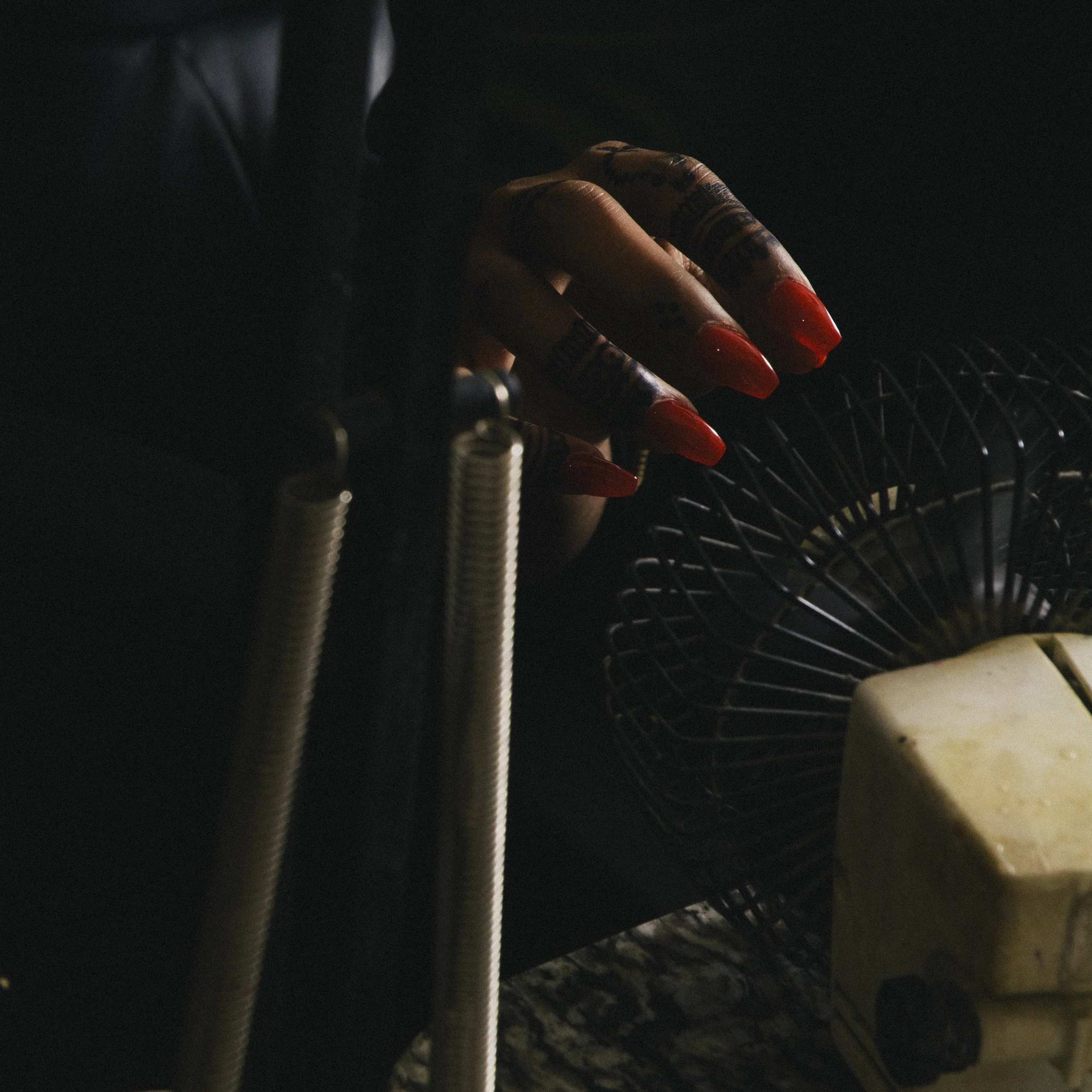 A person's hand with long red nails and tattoos on fingers, near a small portable fan and a yellowish device, in a dark environment.