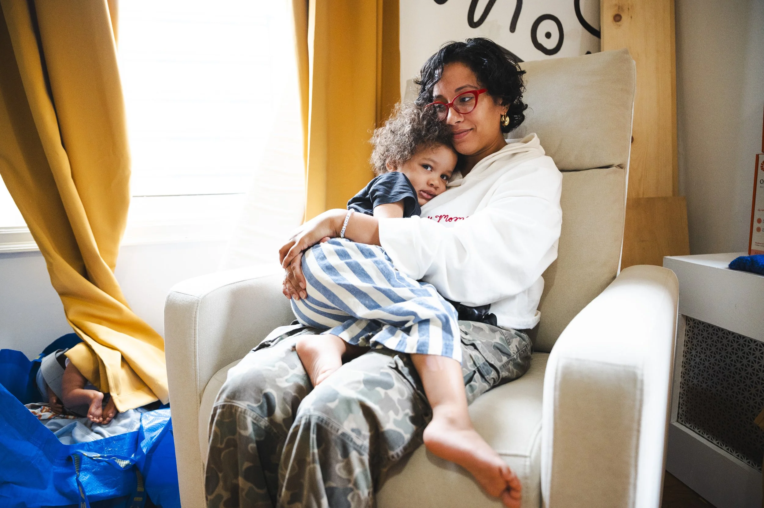 A woman wearing glasses and camouflage pants sits in a beige armchair, holding a small child with curly hair, wearing striped pajama pants, in her lap, in a room with yellow curtains and natural light.