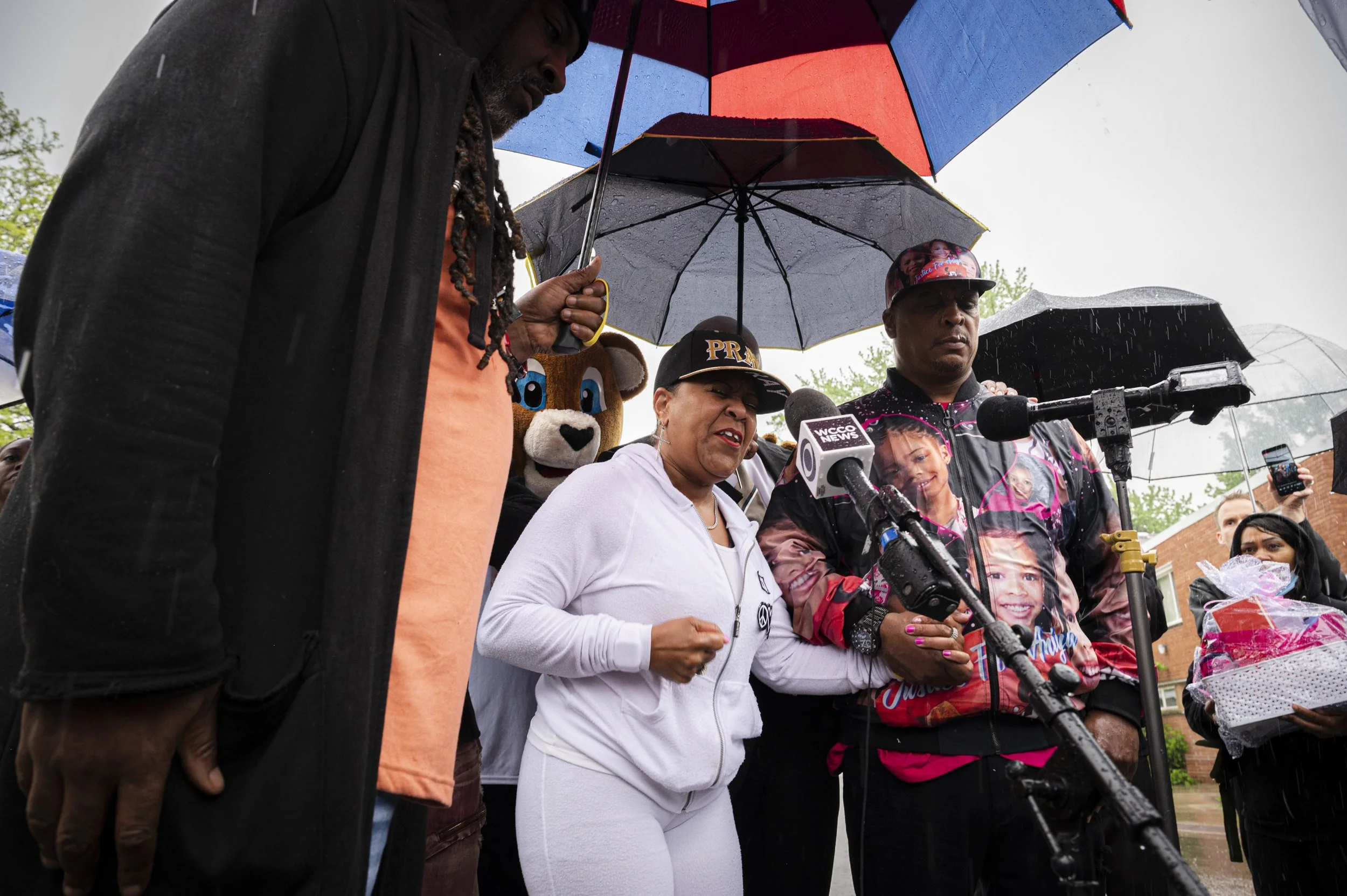 Group of people standing outside in the rain, holding umbrellas, involved in a press conference or speech. One woman is speaking into multiple microphones, and some individuals are wearing custom and casual clothing, including a person in a lion masc