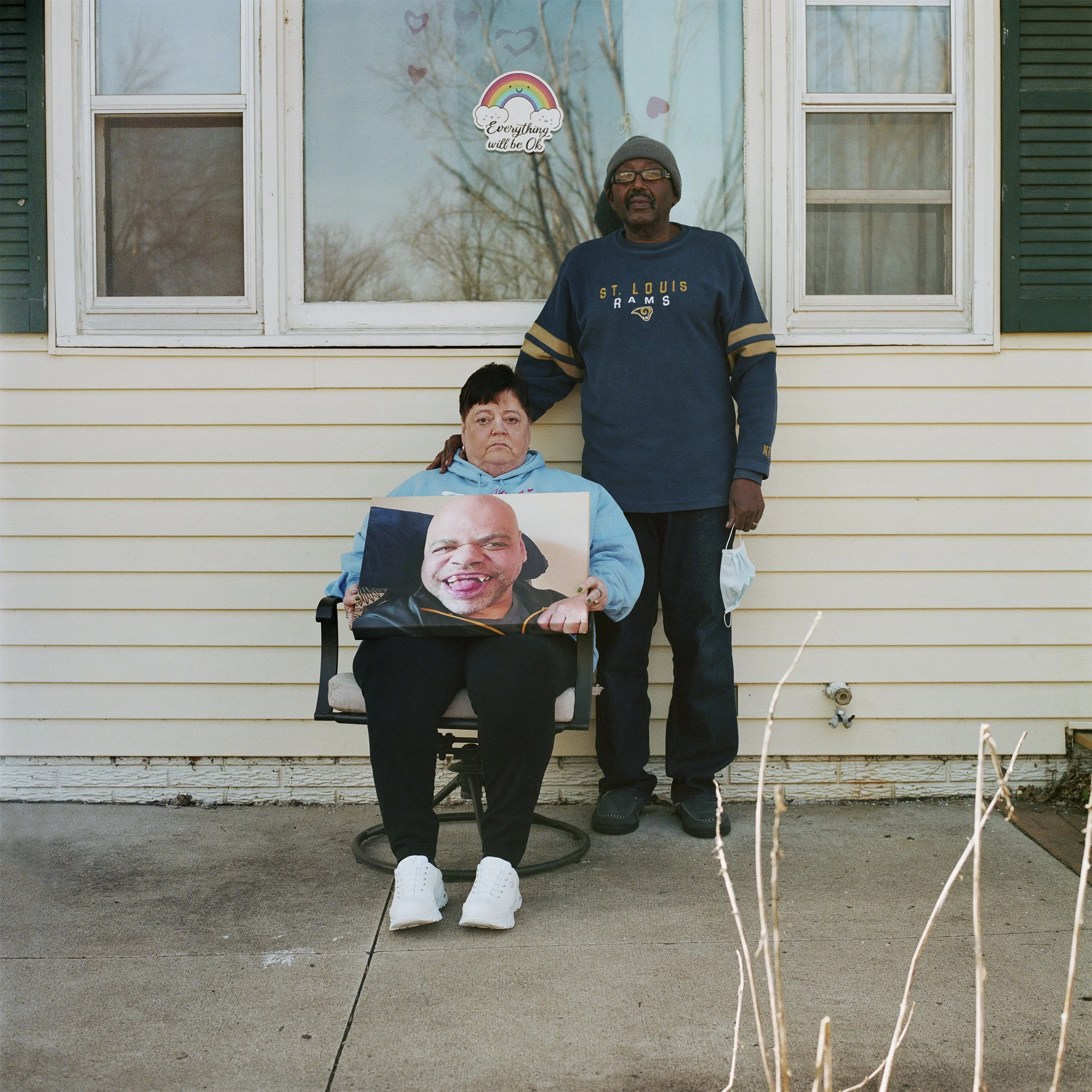 A woman sitting on a chair holding a large photograph of a smiling man with facial hair and a shaved head. Standing beside her is a man in a dark sweatshirt with 'St. Louis Rams' written on it, holding a disposable face mask, outside a yellow house with green shutters and two windows. A sticker on the window says 'Everything will be Ok' with a rainbow and clouds.