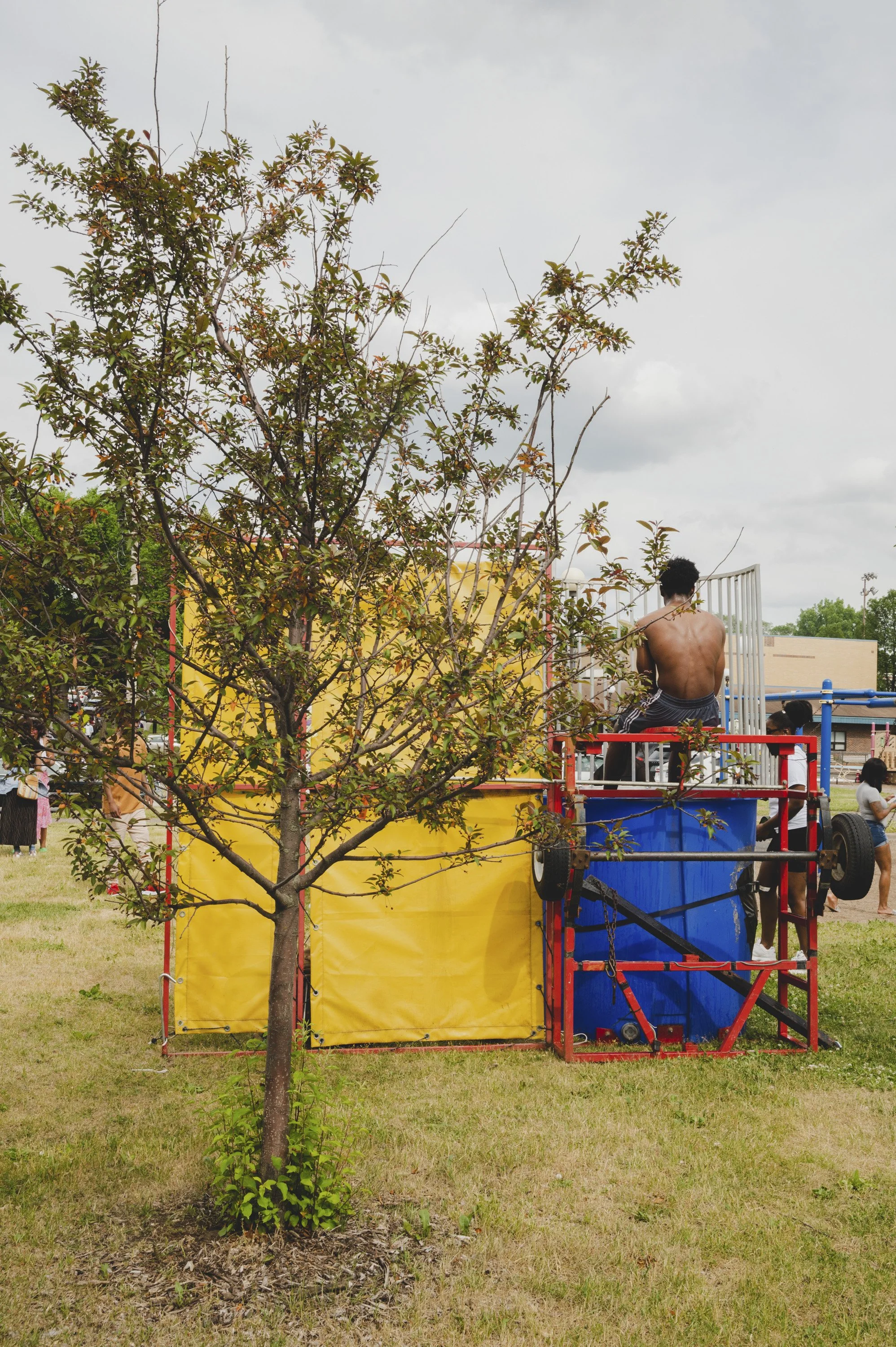Children playing at a dunk tank at an outdoor Juneteenth event in Minneapolis, MN, with a small tree in the foreground and people in the background.