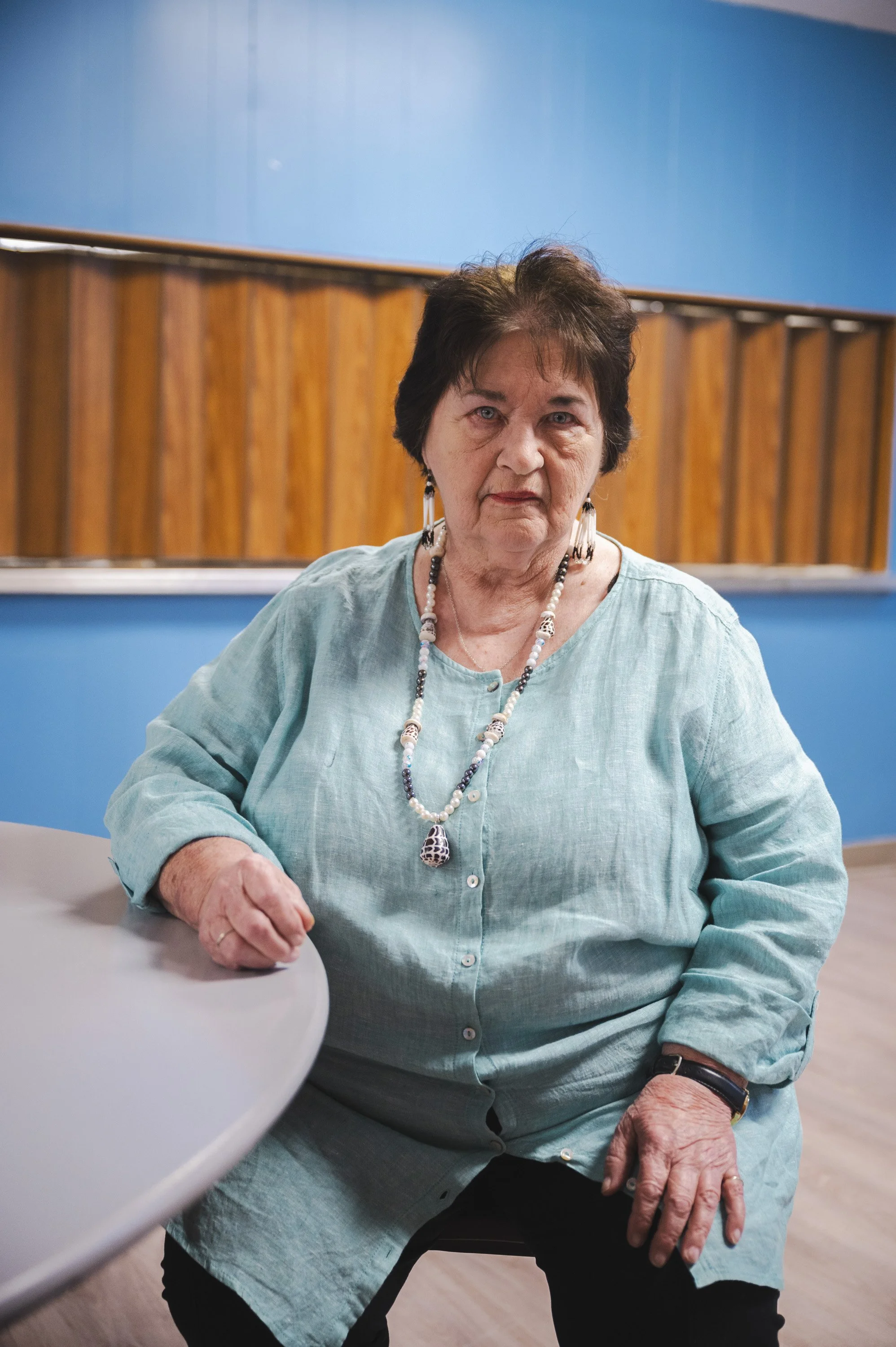 An elderly woman with short dark hair wearing a light blue shirt, decorated with necklaces, sitting at a table in a room with a blue wall and wooden panels.