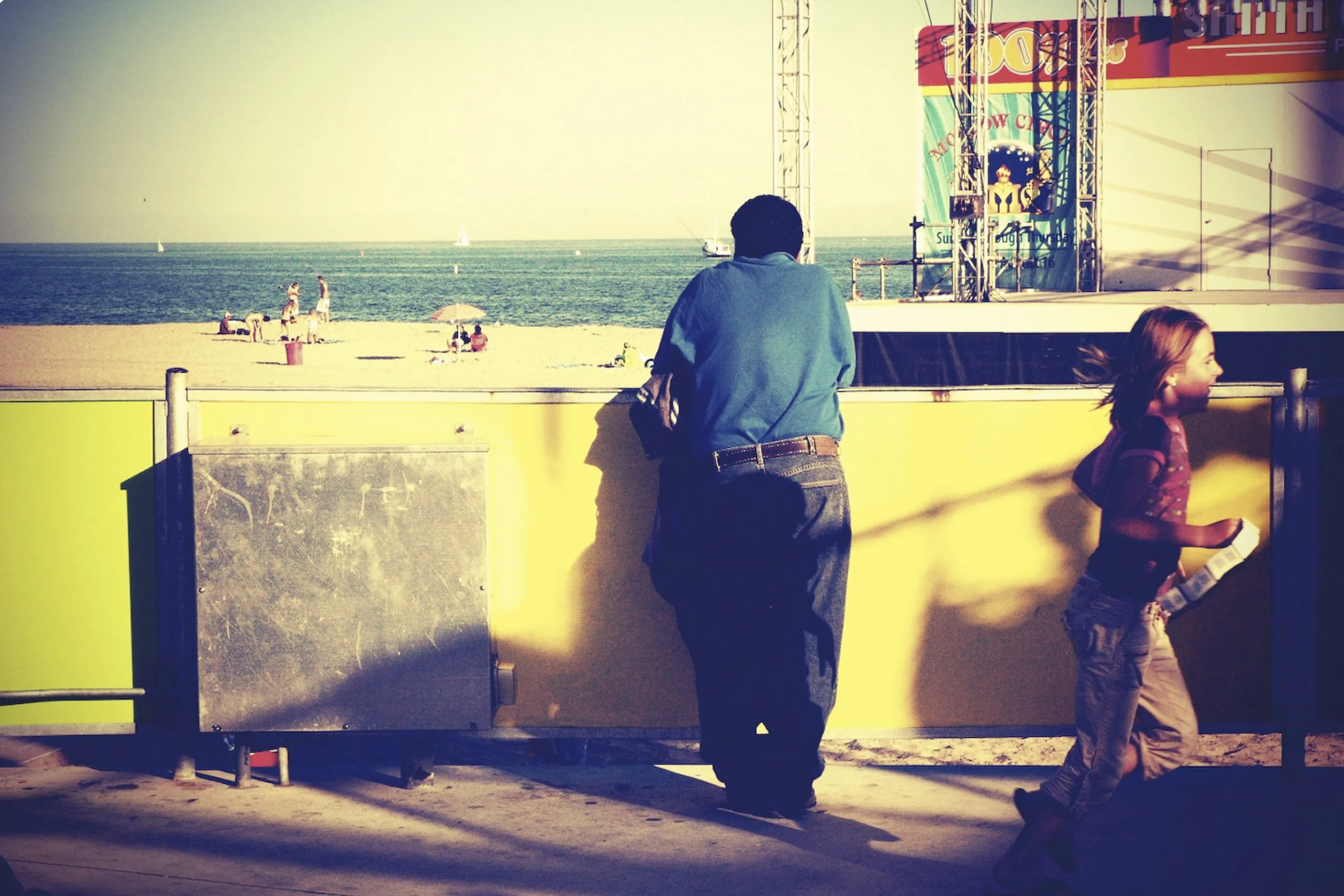 Man and girl near a yellow barrier watching a beach scene with people, sand, and boats in the ocean. An amusement park ride with colorful signage is partially visible to the right.