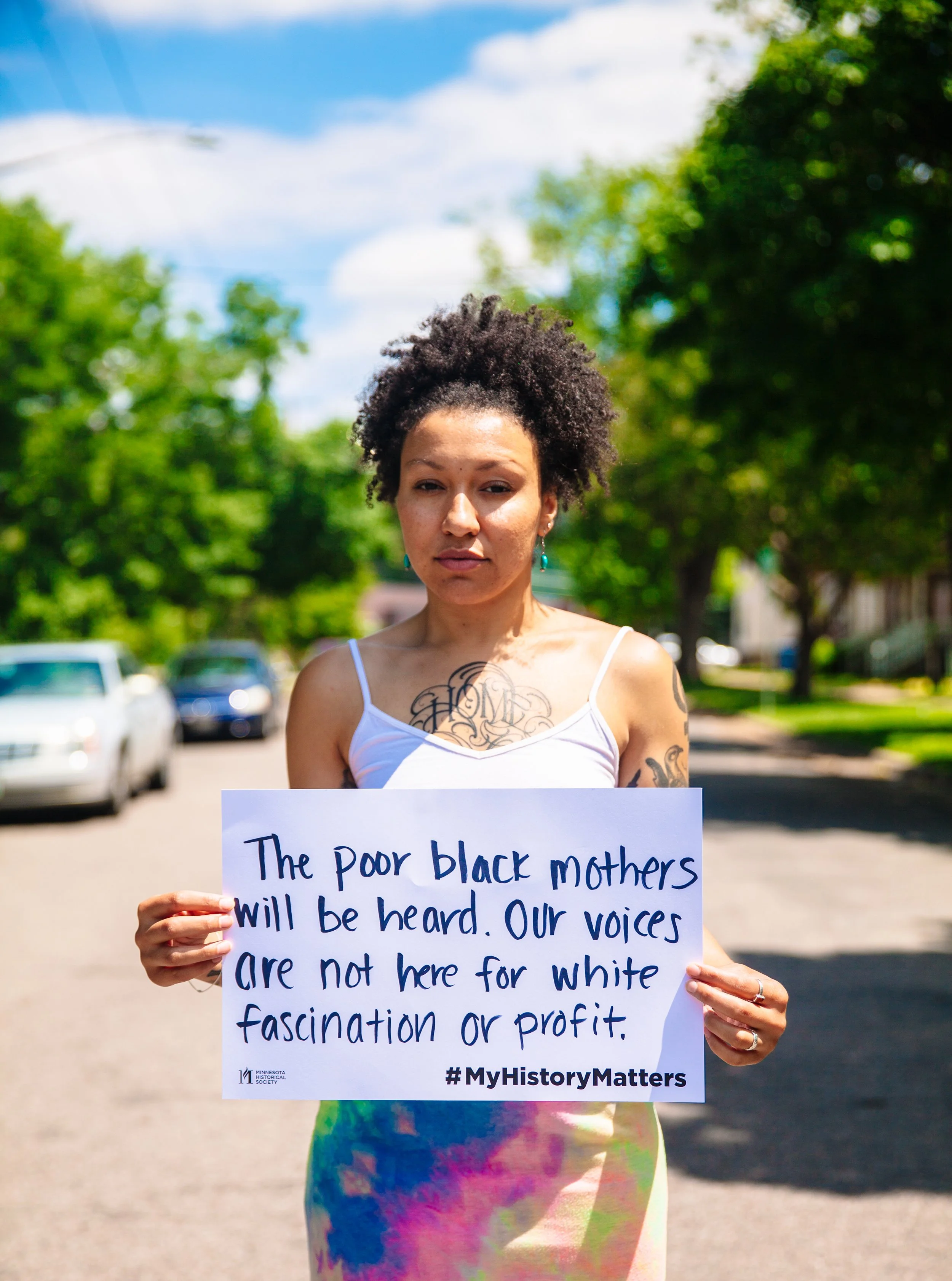 A woman holding a sign during a protest on a tree-lined street. The sign reads: "The poor black mothers will be heard. Our voices are not here for white fascination or profit. #MyHistoryMatters."