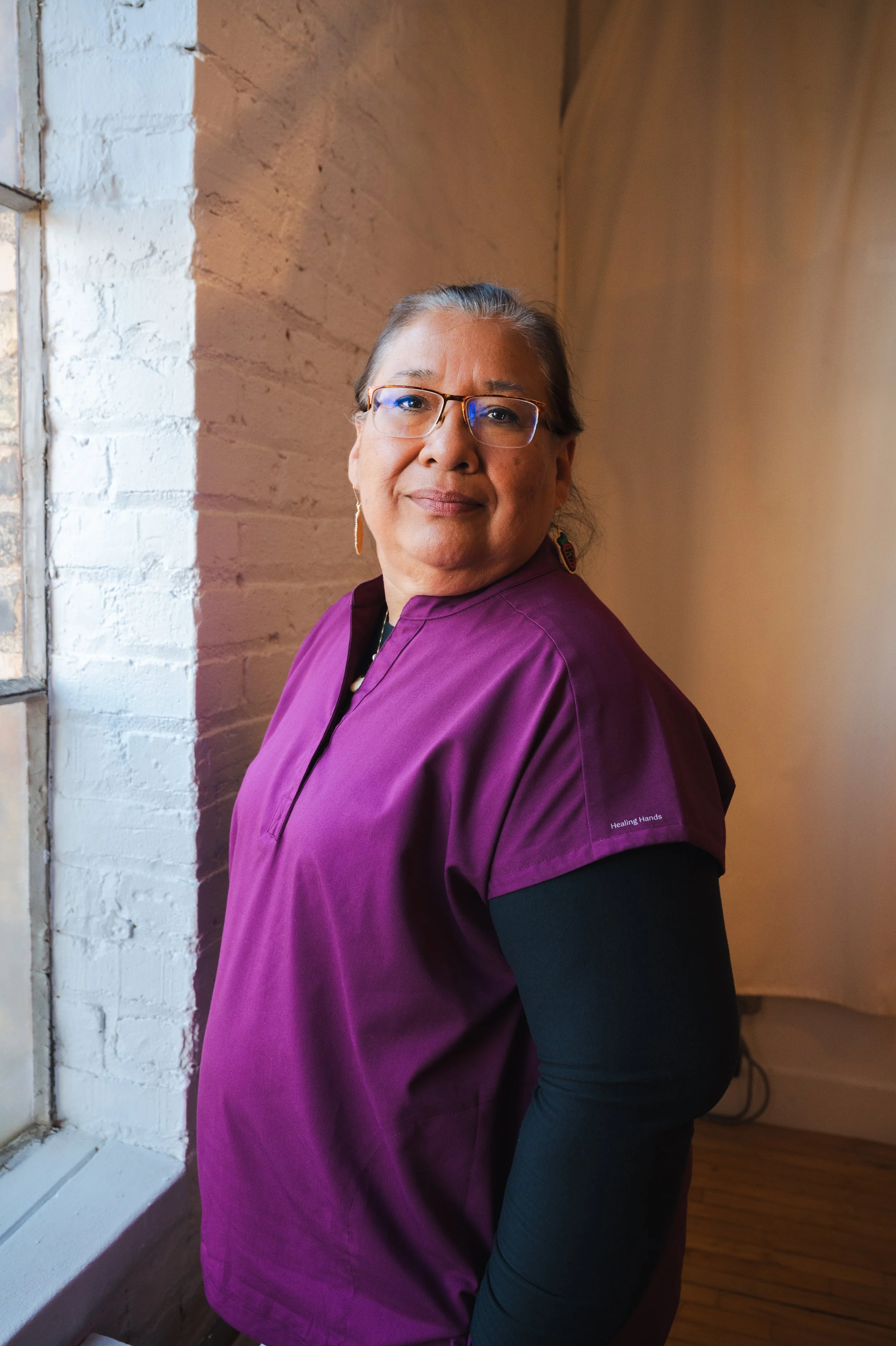 A woman wearing glasses and a purple scrub top stands near a window with white brick wall behind her, smiling softly.
