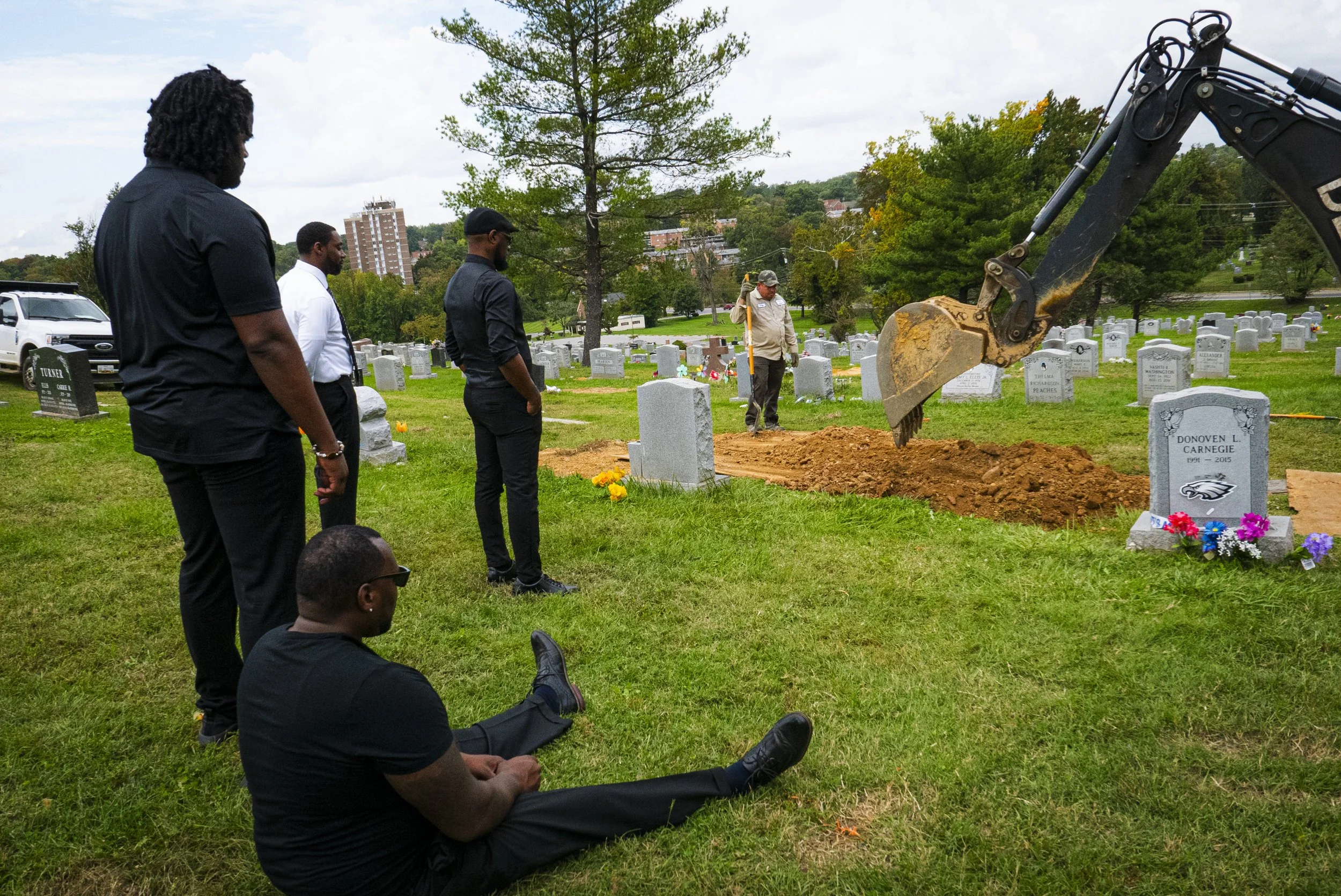 A group of six men in black and white clothing paying respects at a gravesite in a cemetery as an excavator digs a fresh grave. One man is seated on the grass nearby.