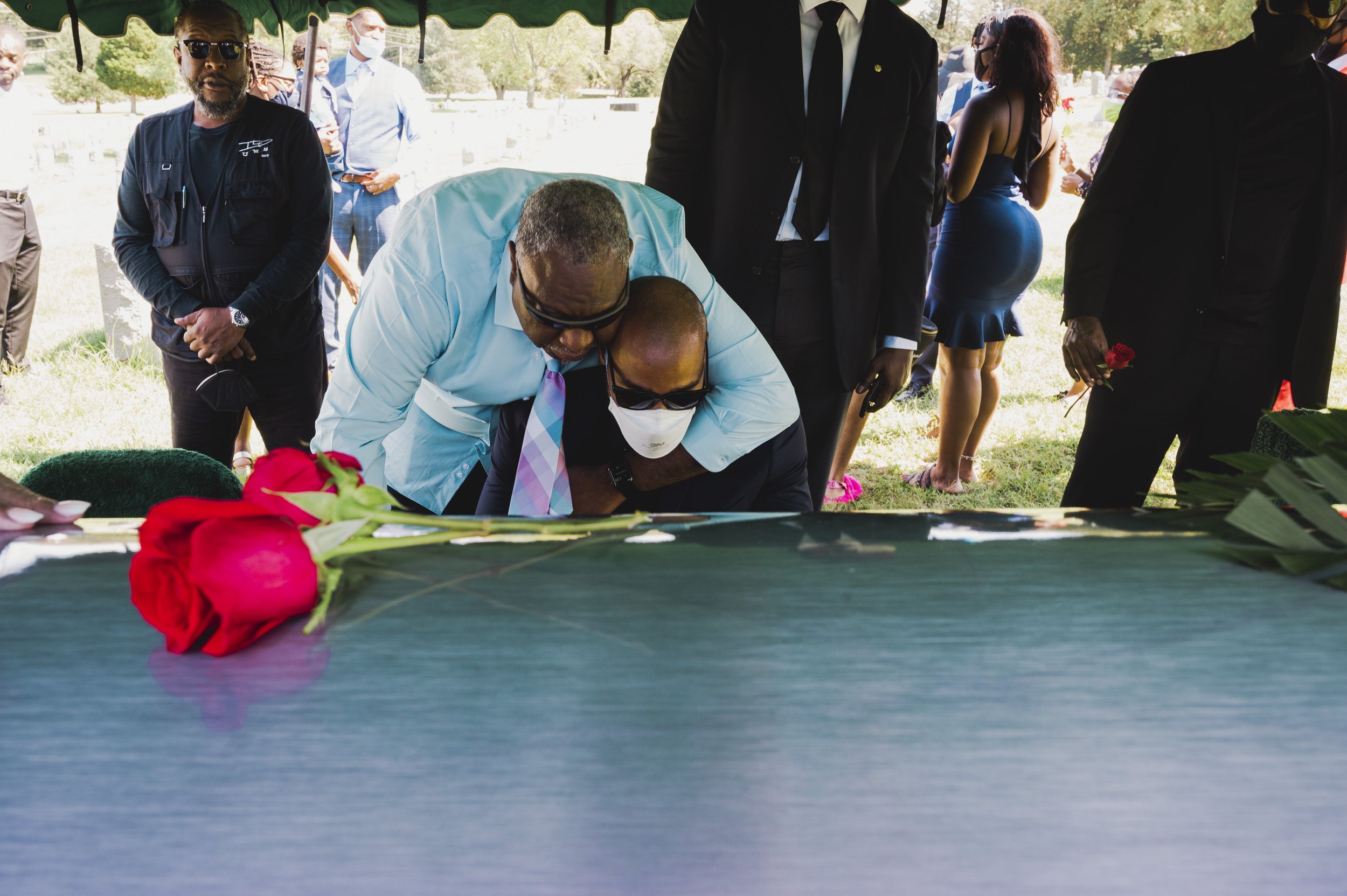 People attending a funeral outdoors, some dressed in formal black suits, with one person placing a hand on a person's back who is wearing a face mask and sunglasses, at a casket with red roses on it.