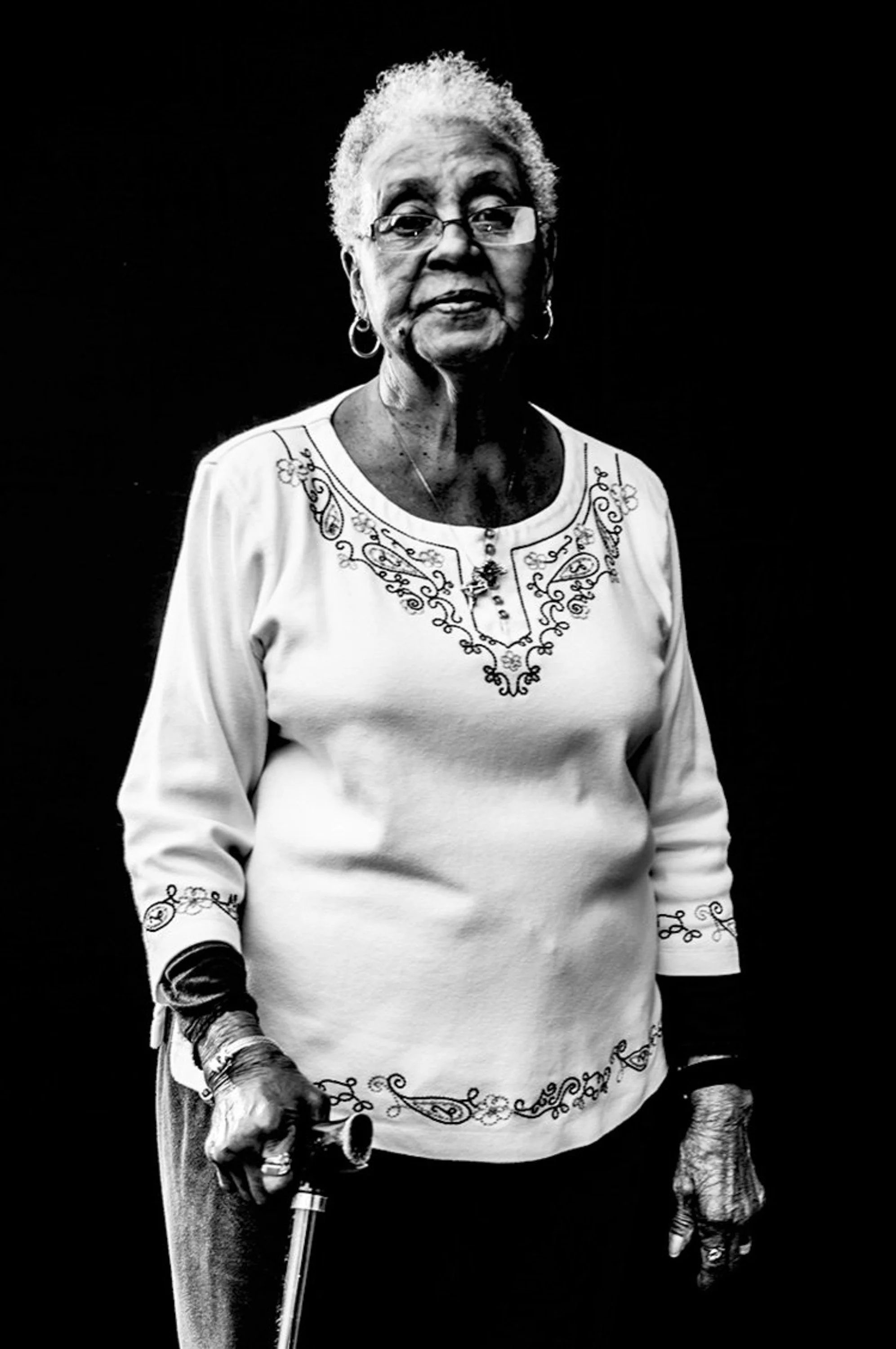 Black and white photo of an elderly woman with short curly hair, wearing glasses, a decorated blouse, and holding a cane.