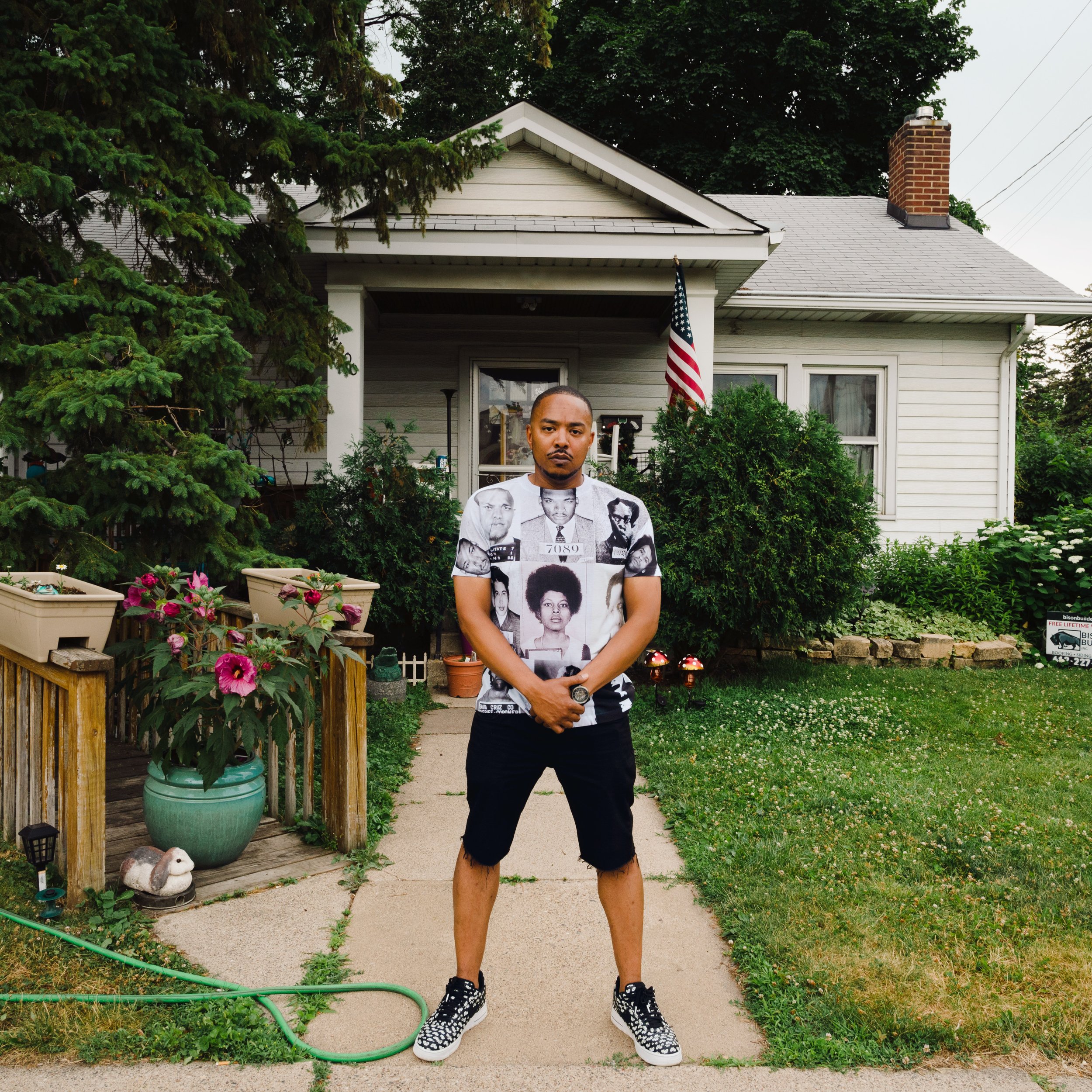 A man standing on a sidewalk in front of a house with a garden, wearing a graphic t-shirt with black-and-white images of various people, black shorts, and sneakers.