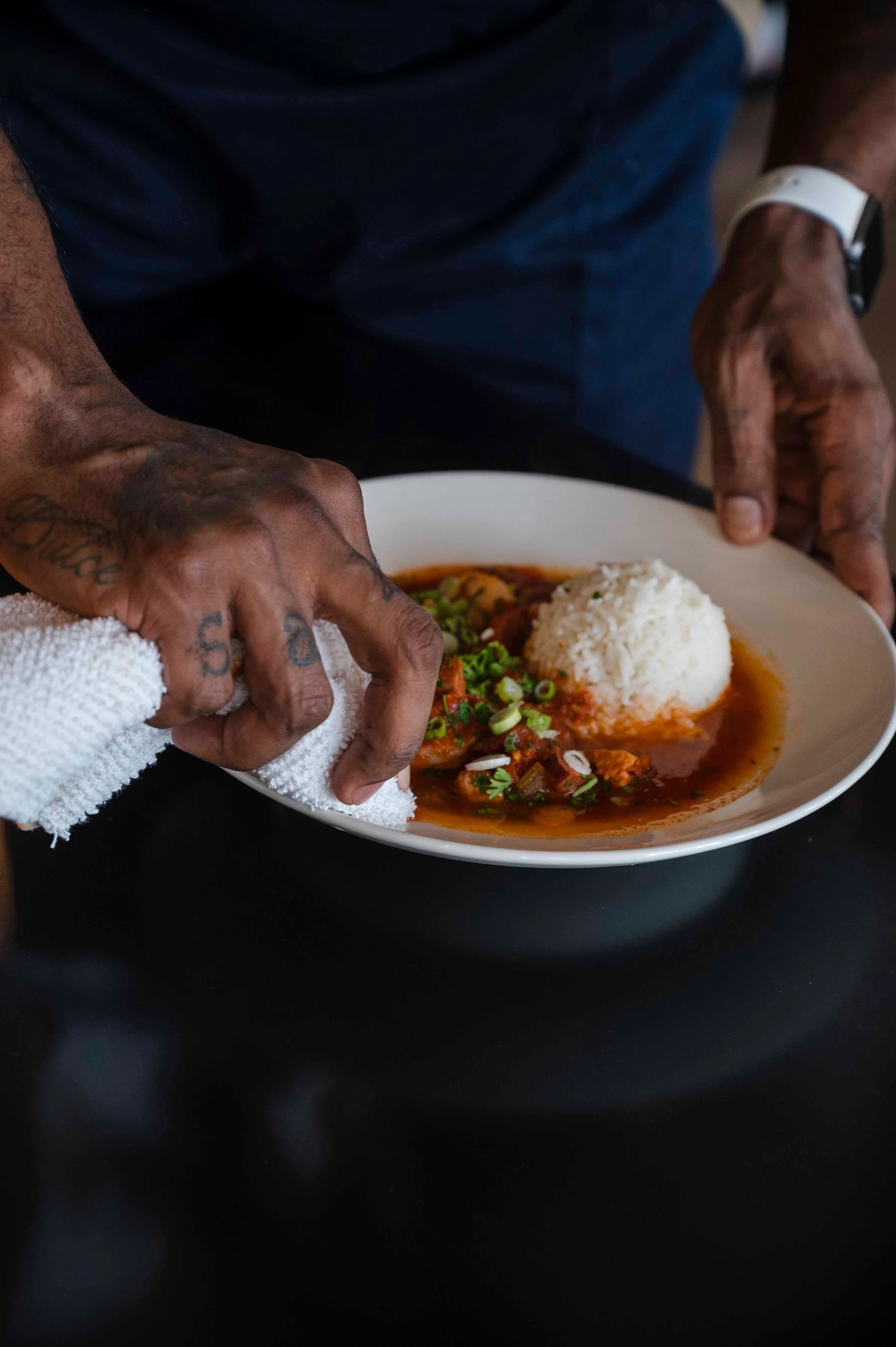 A person with tattooed hands serves a bowl of beef stew with rice garnished with green onions and cilantro.