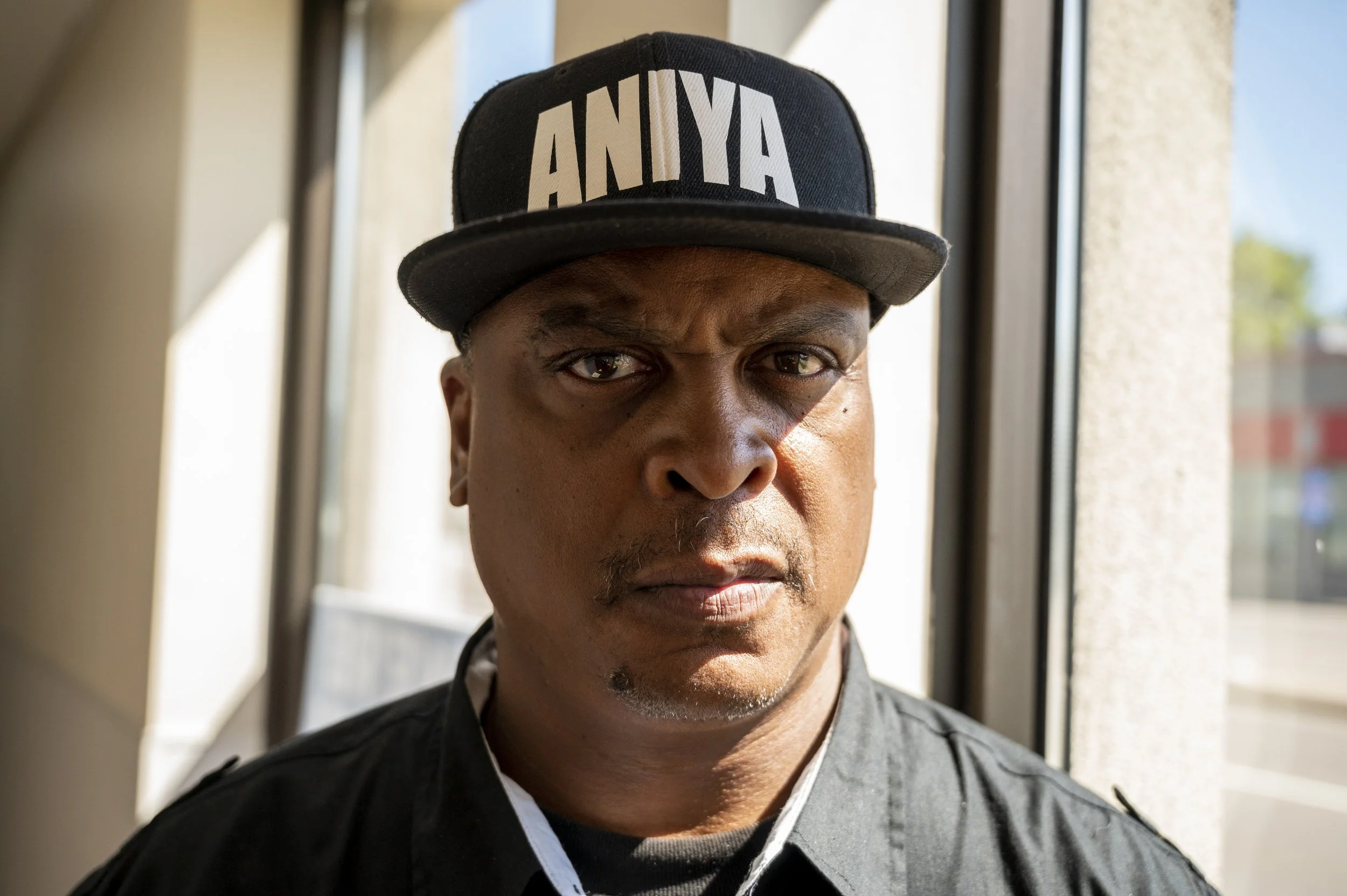 Close-up of a serious Black man wearing a black cap with white letters spelling 'ANIYA' and a black jacket, standing indoors near a window with daylight.