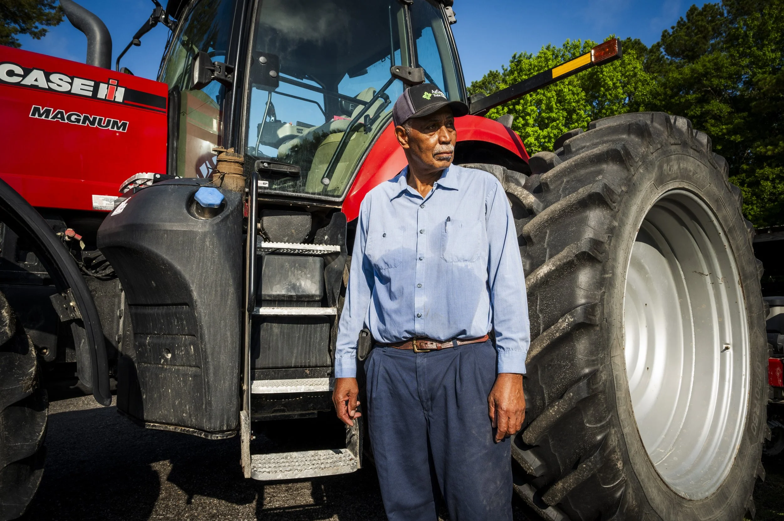 A man standing beside a large red tractor with black tires, wearing a light blue button-up shirt, dark pants, and a baseball cap, outdoors on a sunny day with green trees in the background.
