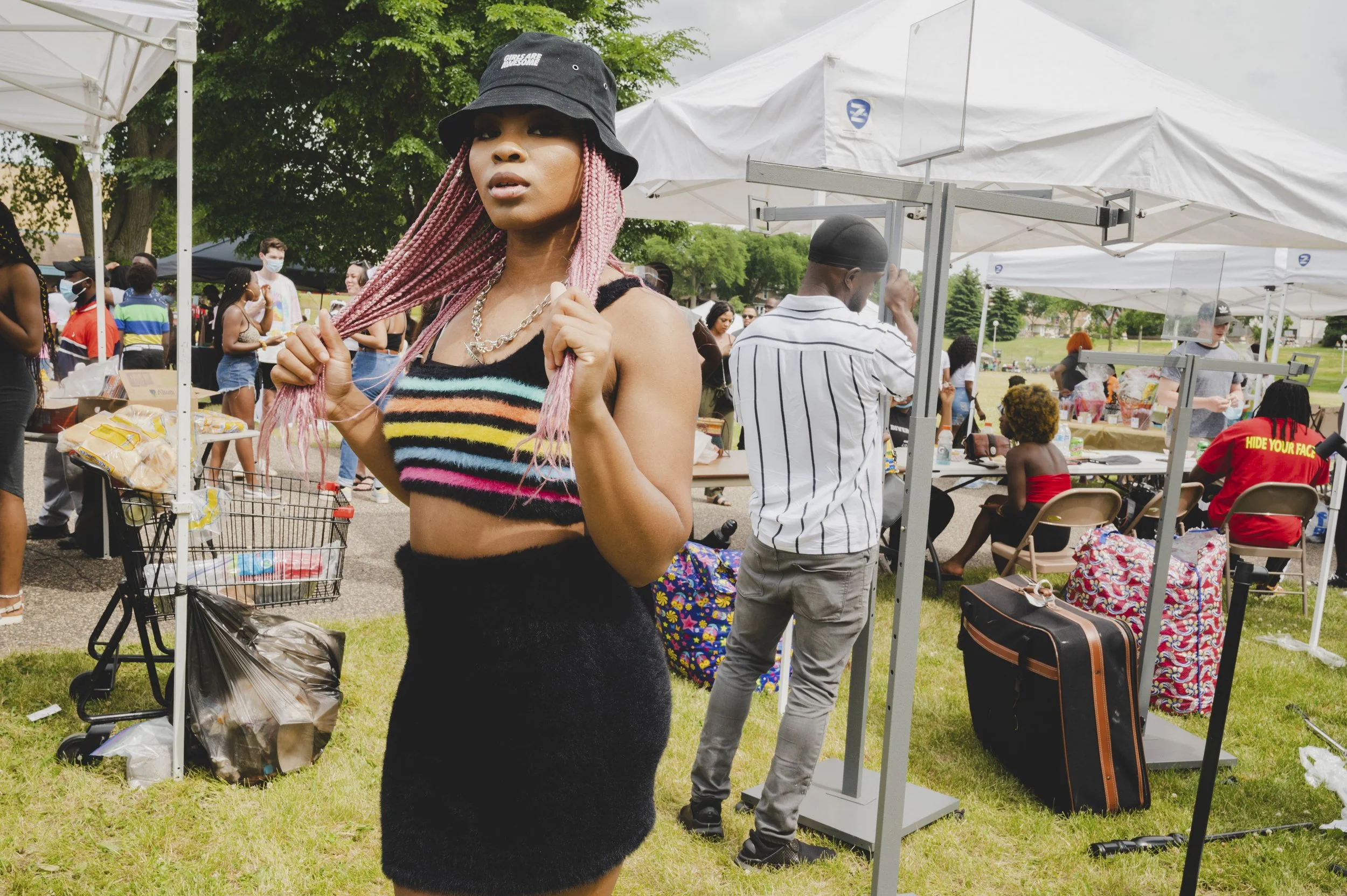 A woman with pink braided hair wearing a black bucket hat and colorful striped crop top in a crowded outdoor park or fair with white tents and people walking and sitting around.