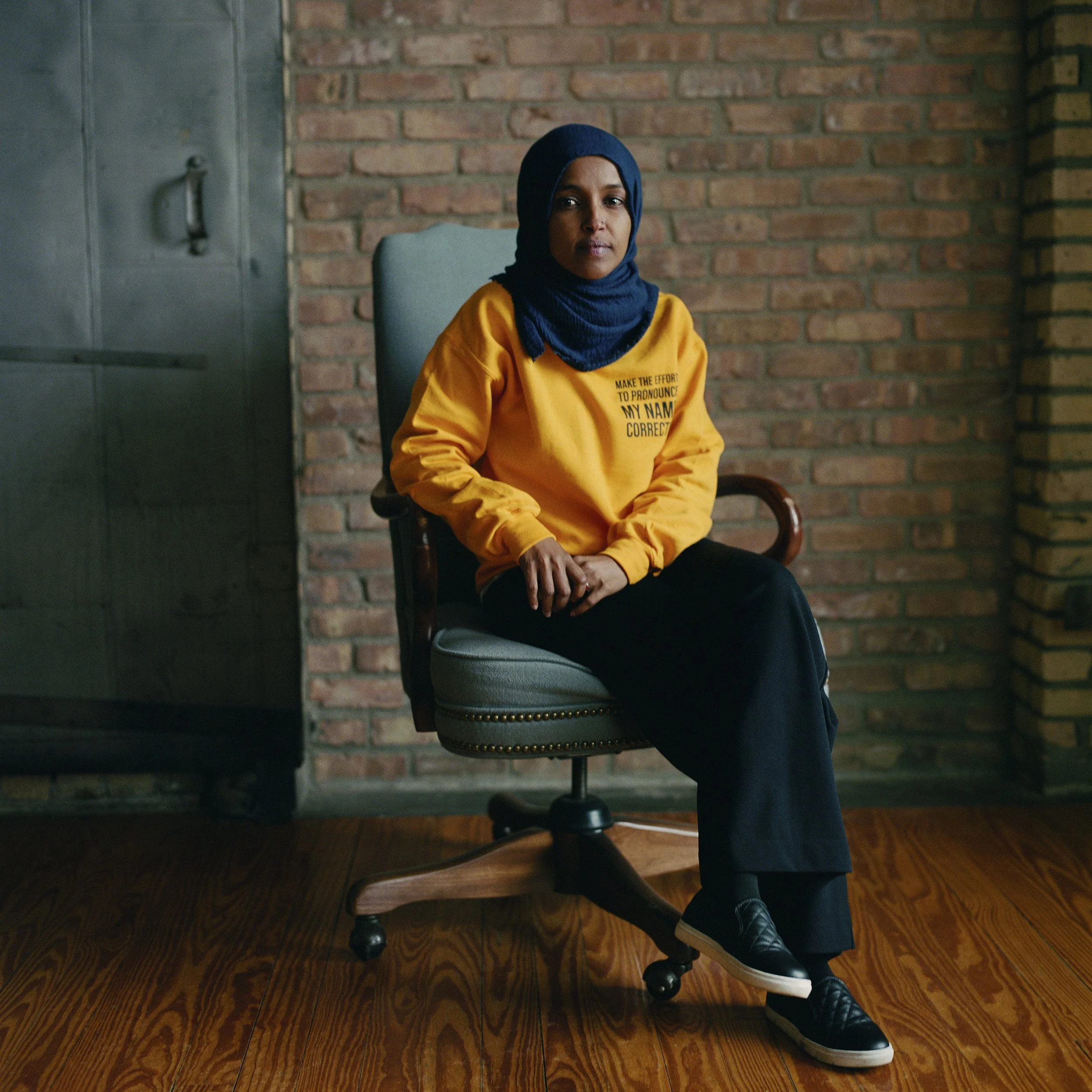 A woman wearing a blue hijab and a yellow sweatshirt sitting on an office chair in front of a brick wall.