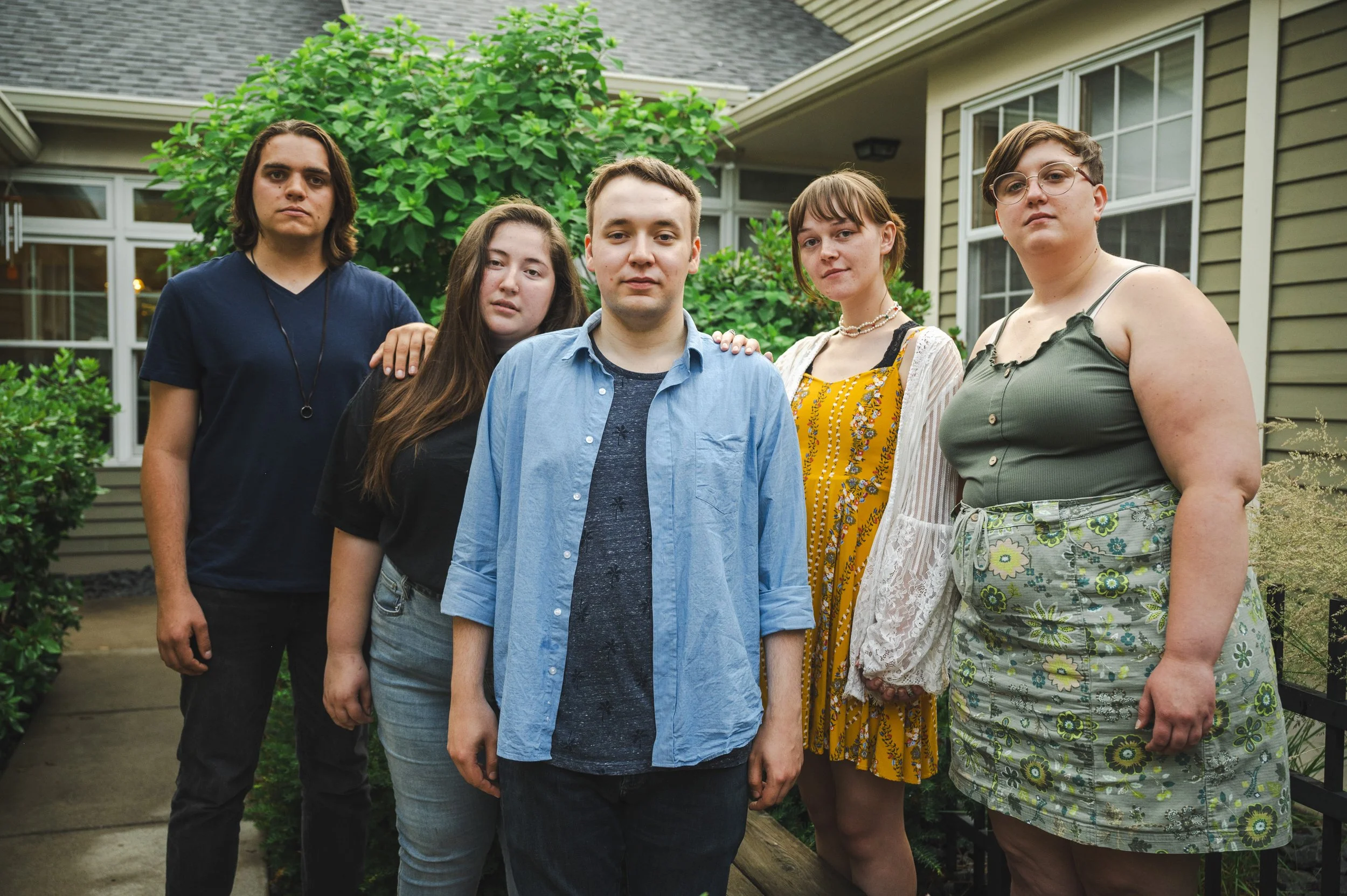 A group of five young adults standing outdoors in front of a house with greenery, looking at the camera with neutral expressions.