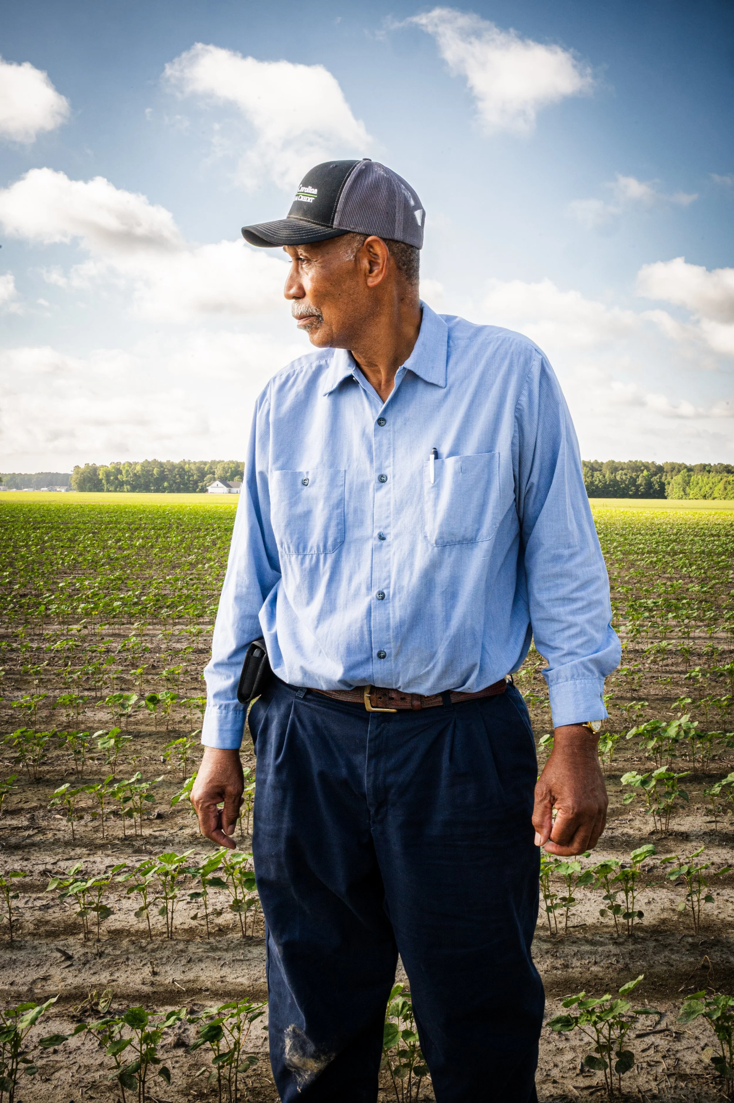 An older man with a mustache wearing a blue button-up shirt, dark pants, and a black cap, standing in a field with small green plants under a blue sky with scattered clouds.