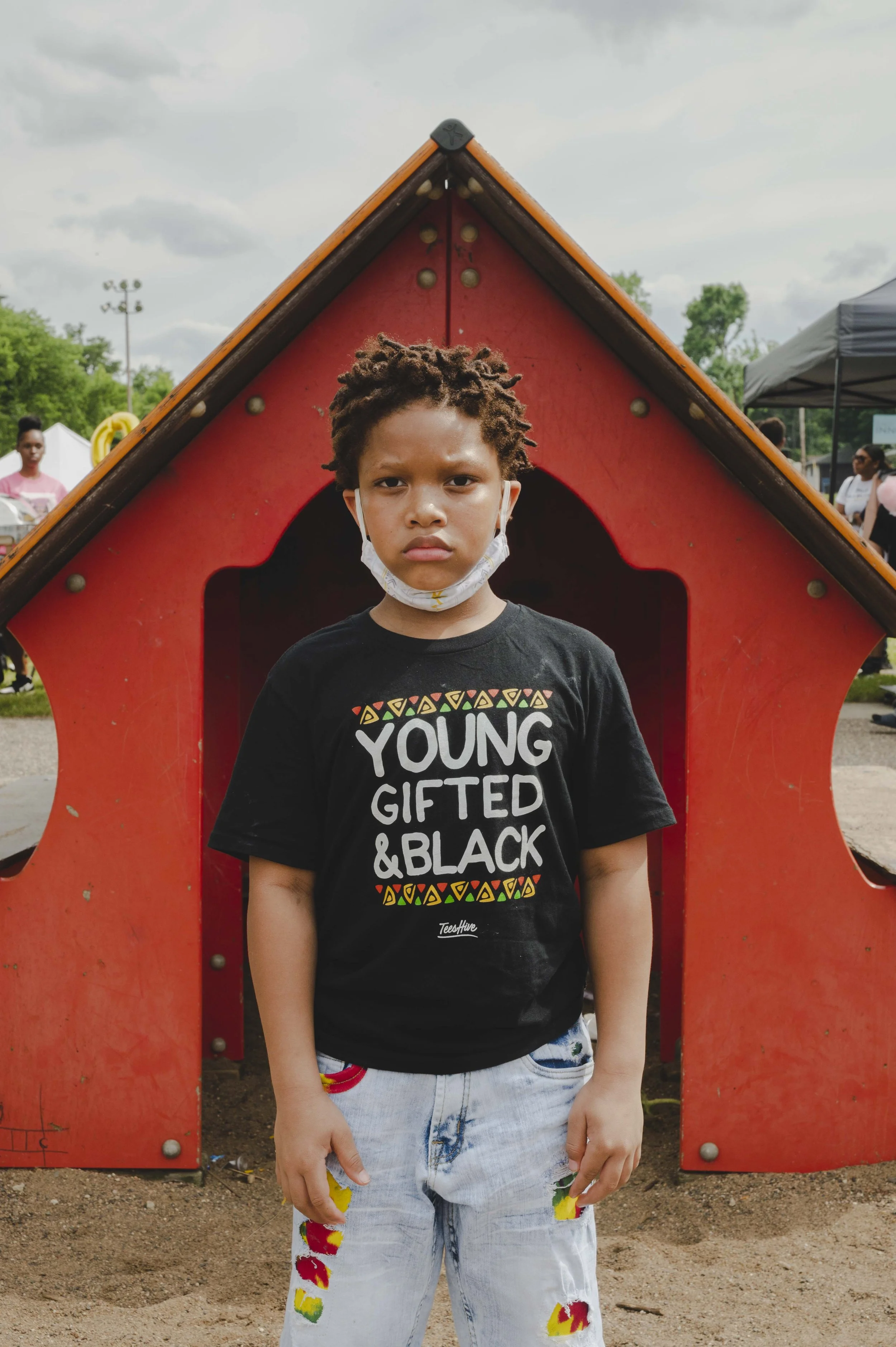 A young boy with dreadlocks standing in front of a red and black play structure at a park. He is wearing a black T-shirt with the words 'YOUNG, GIFTED, & BLACK' printed on it, and light-colored shorts with colorful paint splashes. He has a serious ex