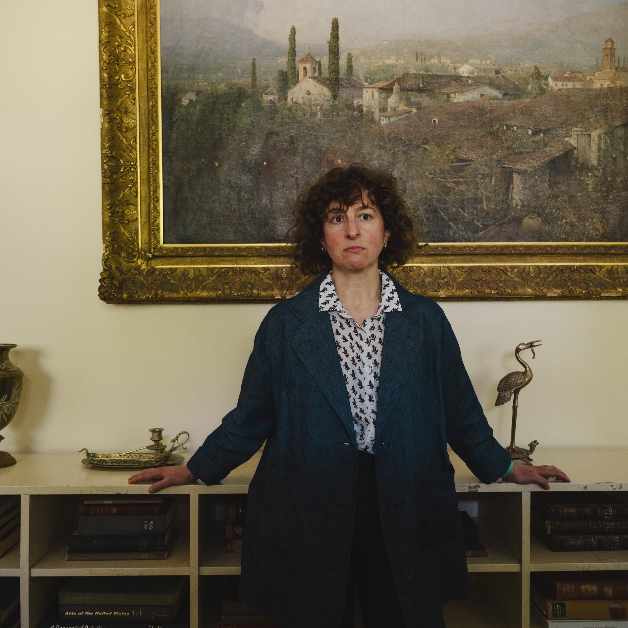 A woman with curly hair standing in front of a large framed landscape painting, resting her hands on a white bookshelf filled with books and decorative items.