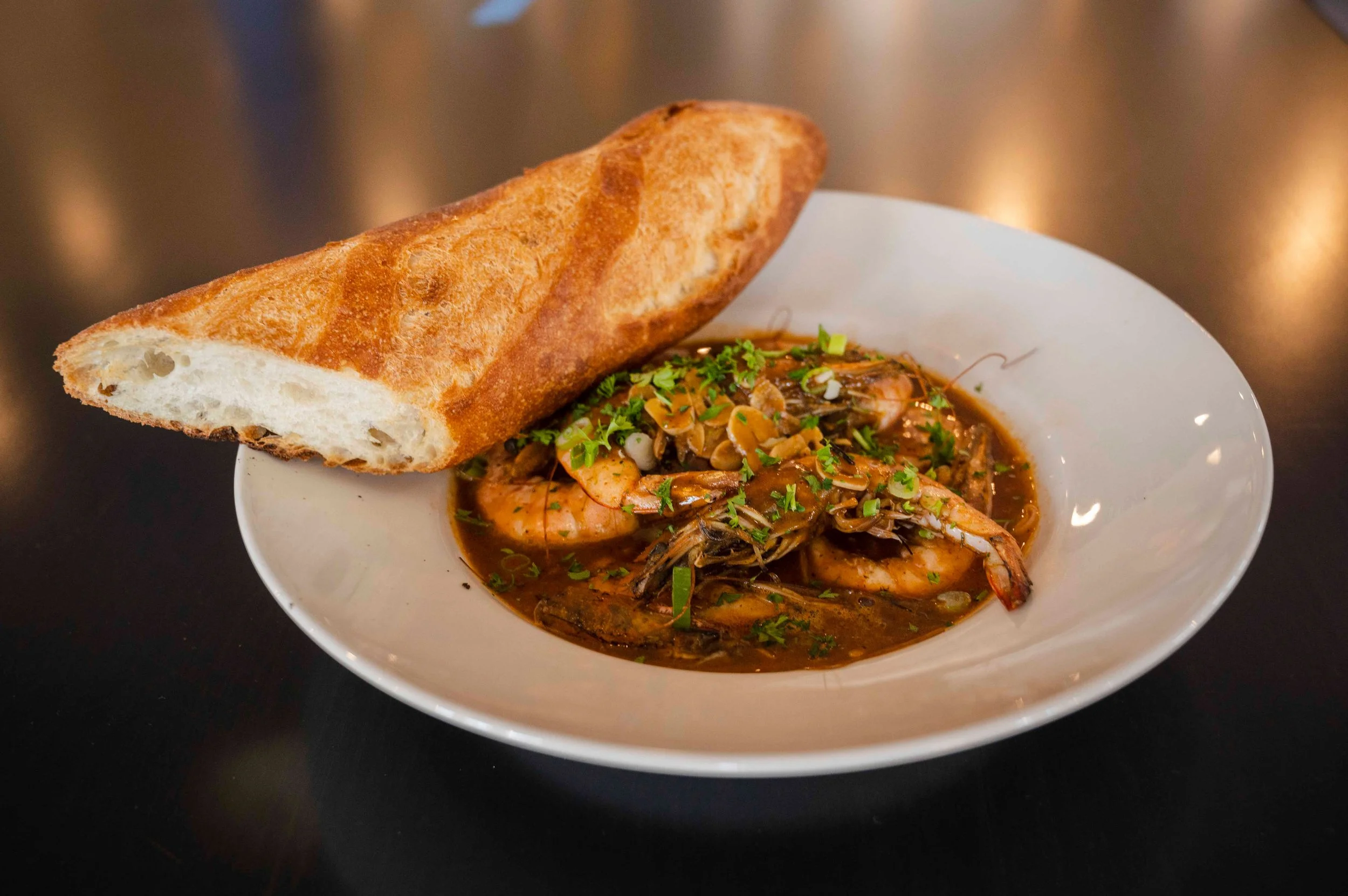 A bowl of shrimp in a dark sauce garnished with chopped herbs, accompanied by a slice of toasted bread on the side.