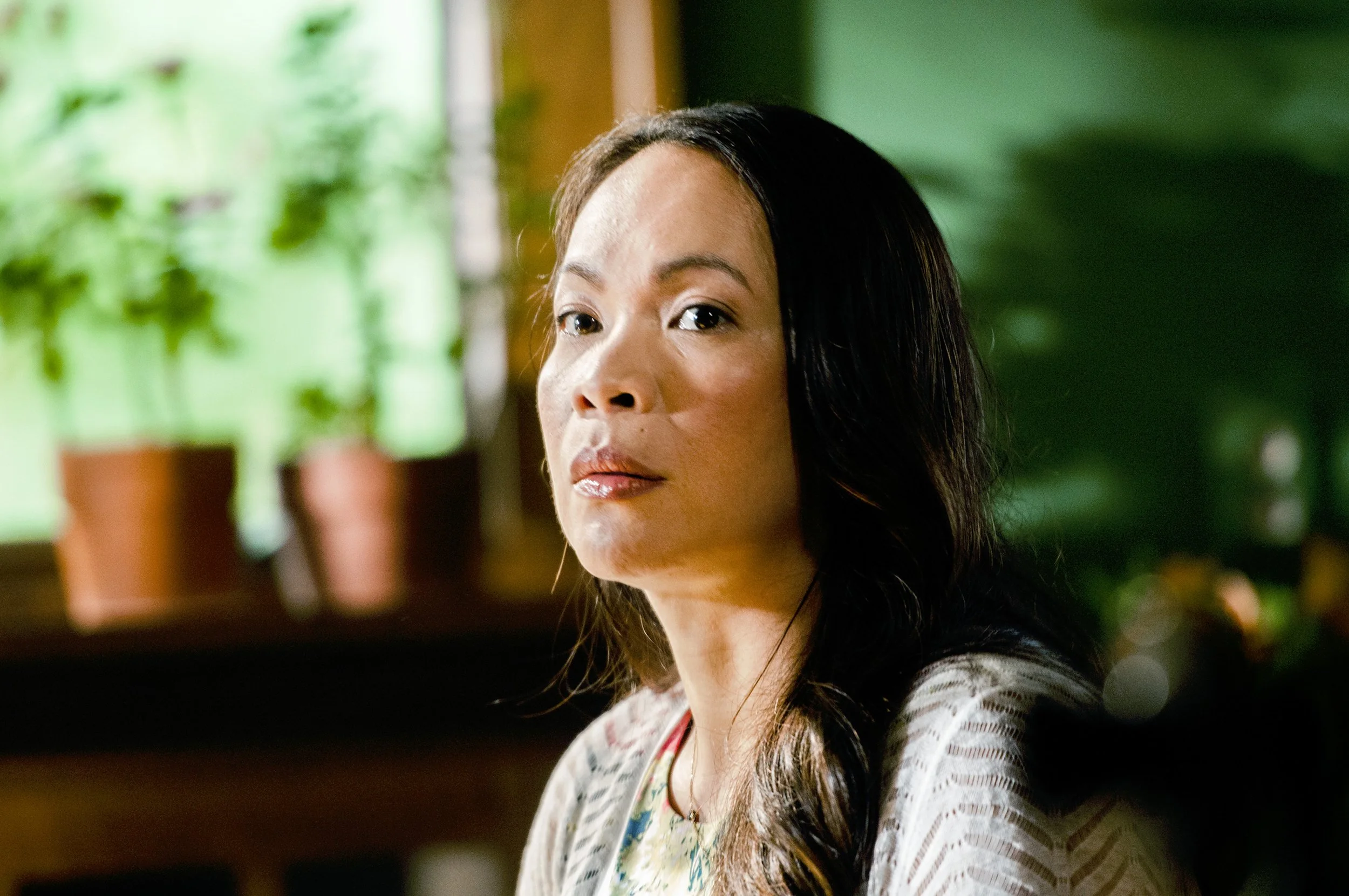 A woman with long dark hair sitting indoors with a green background and some potted plants behind her.