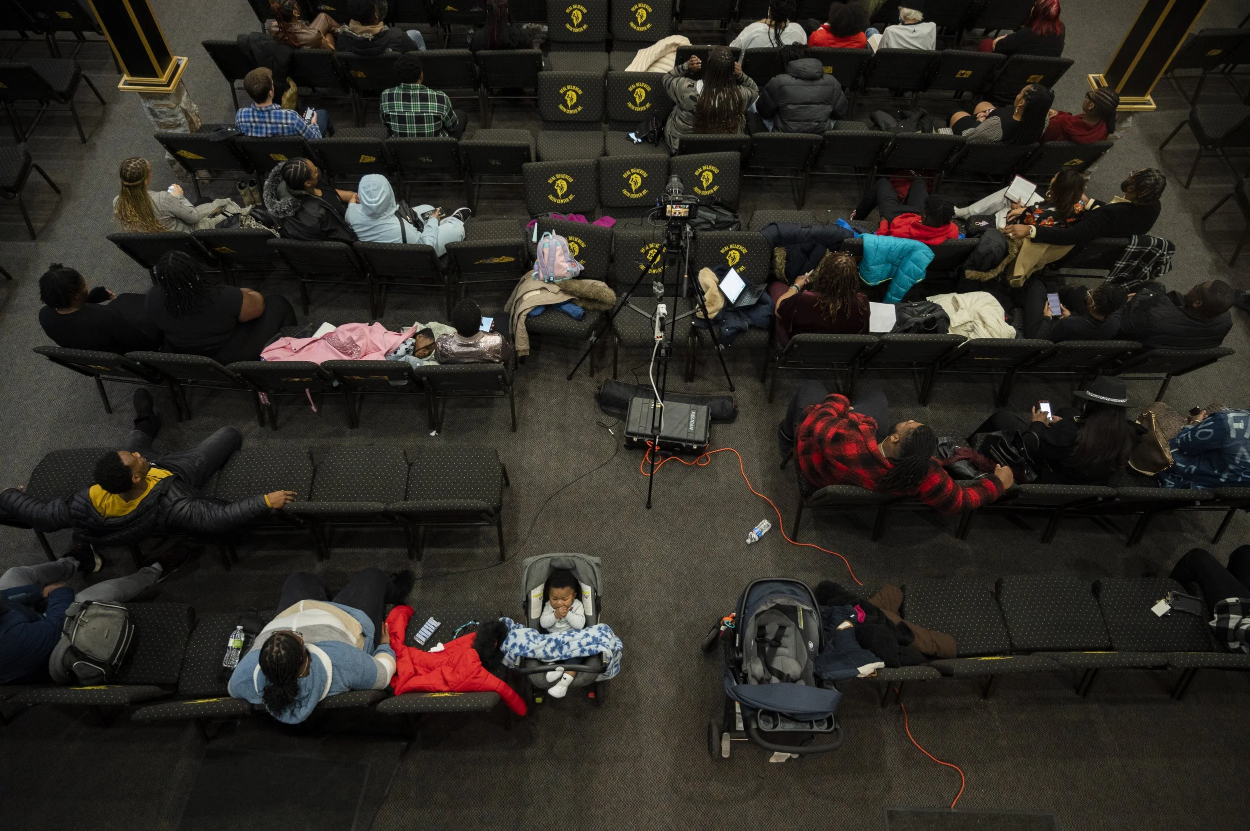 People seated in a church watching the service, some using their phones, with a camera setup in the center.