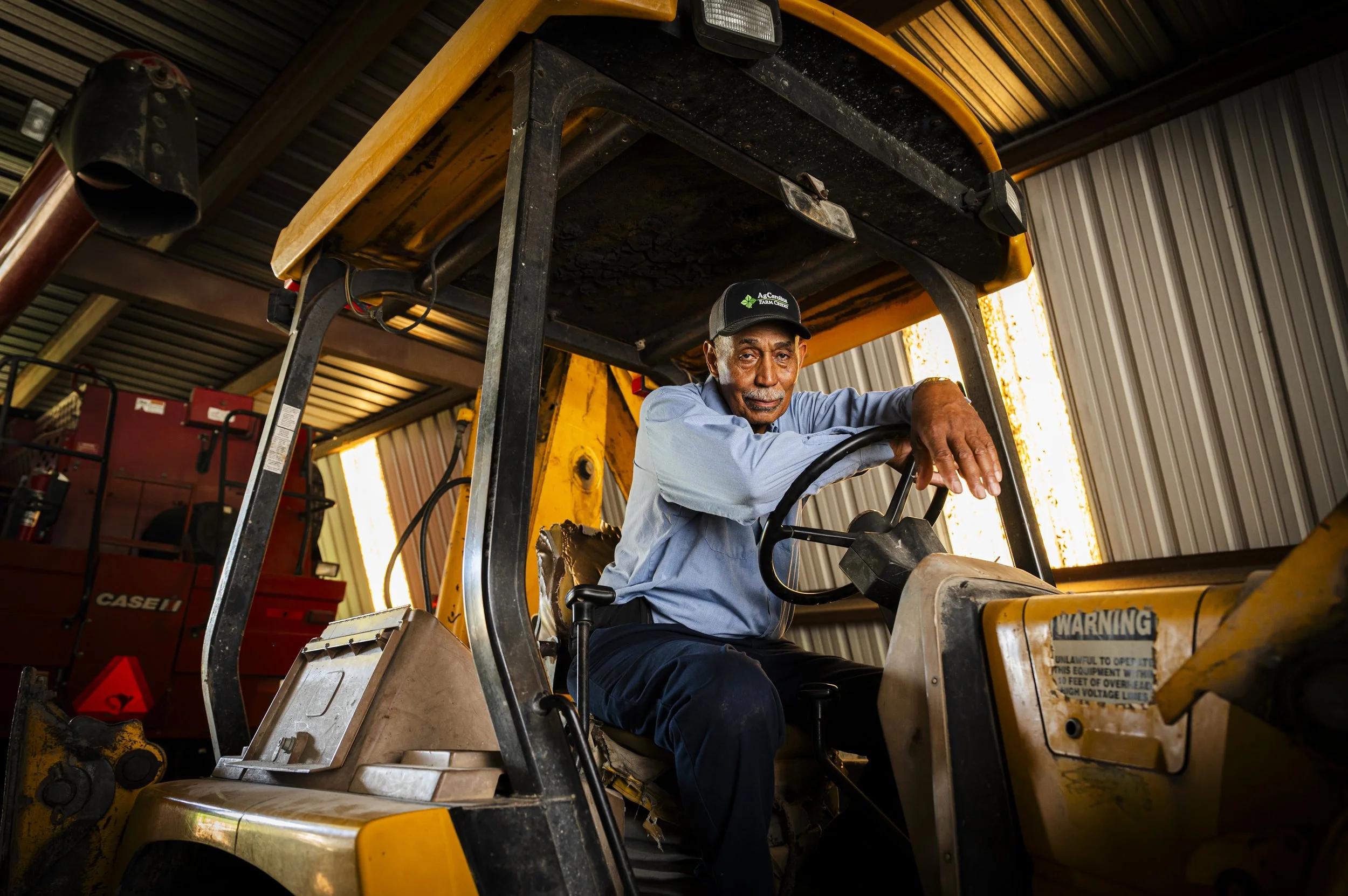 An elderly man sitting in a yellow tractor inside a metal-sided barn.