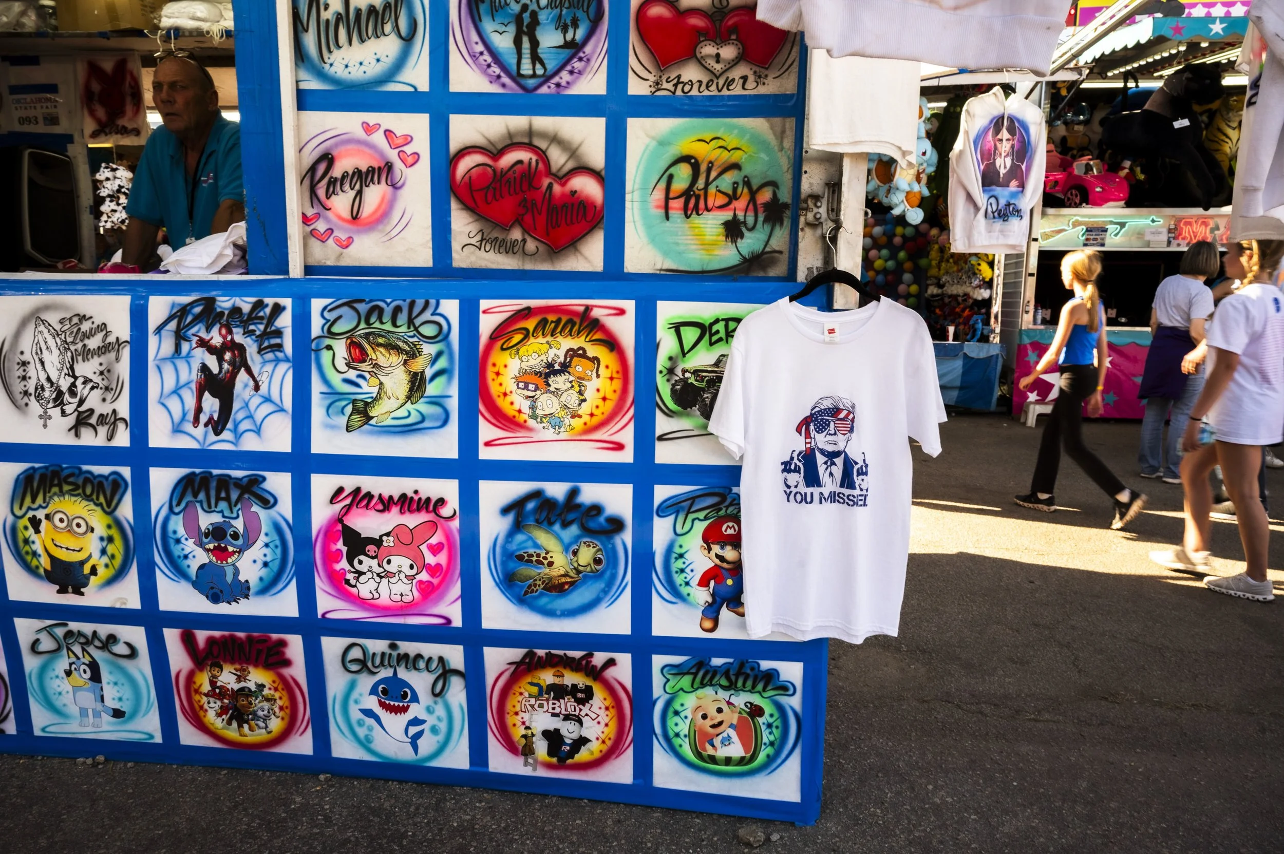 A carnival or fair booth displaying neon-style artwork with personal names, cartoon characters, and custom designs, including a white T-shirt hanging with a graphic of a person with sunglasses and the text "You Missed." People walk past in the backgr