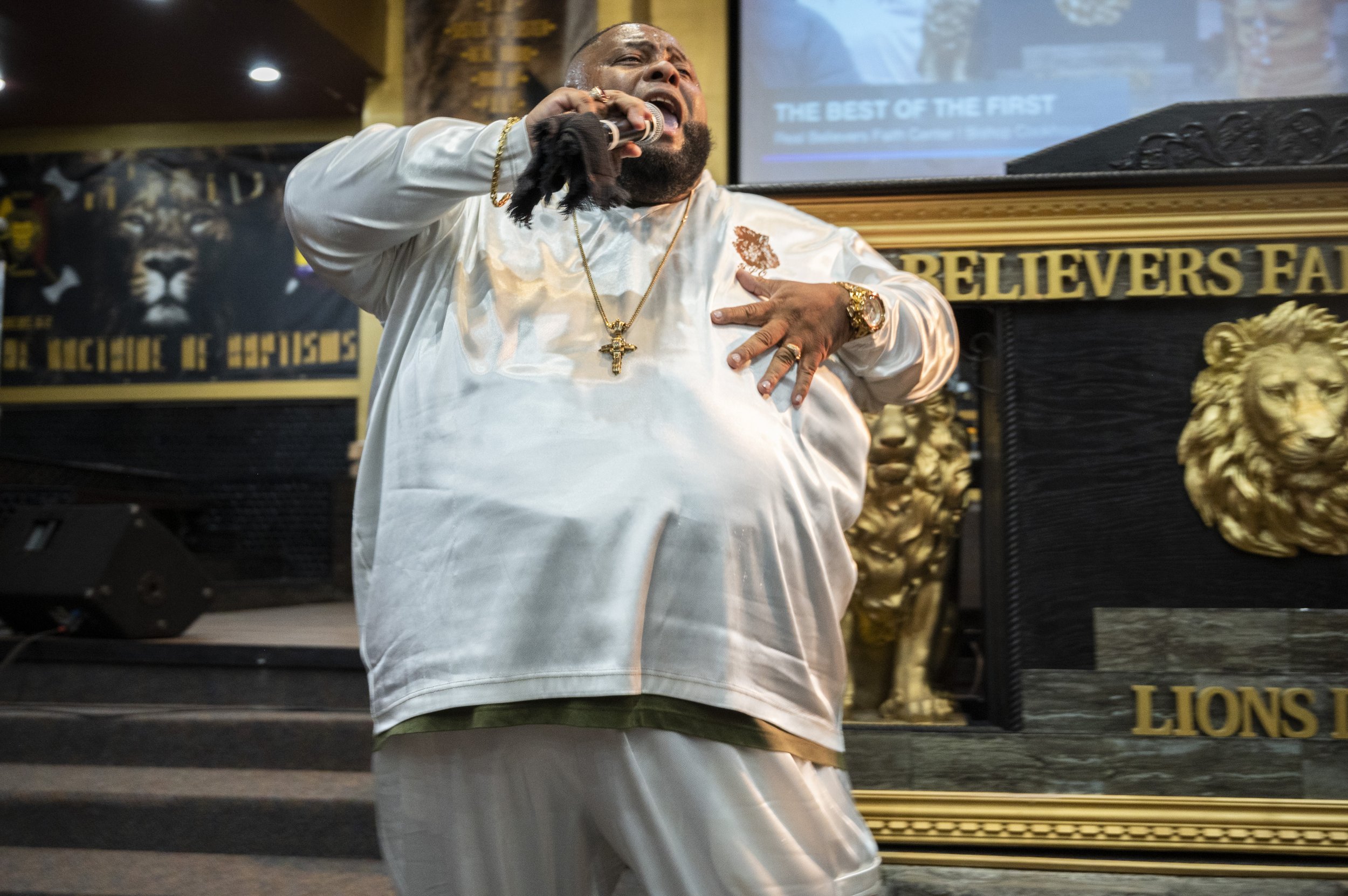 A pastor preaching passionately into a microphone, wearing a white satin shirt and gold jewelry, in front of a lion-themed decorative background in a venue with signs that read 'Believers' and 'Lions'.