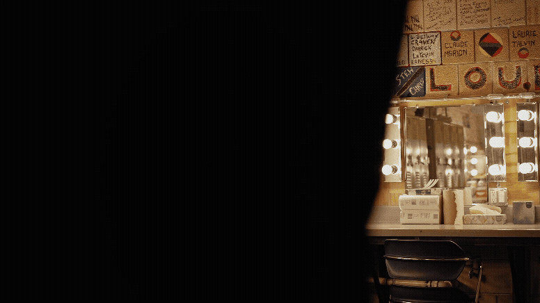 A vanity mirror with bright lights surrounded by various signs and notes on the wall, with a chair and some items on the table below the mirror.