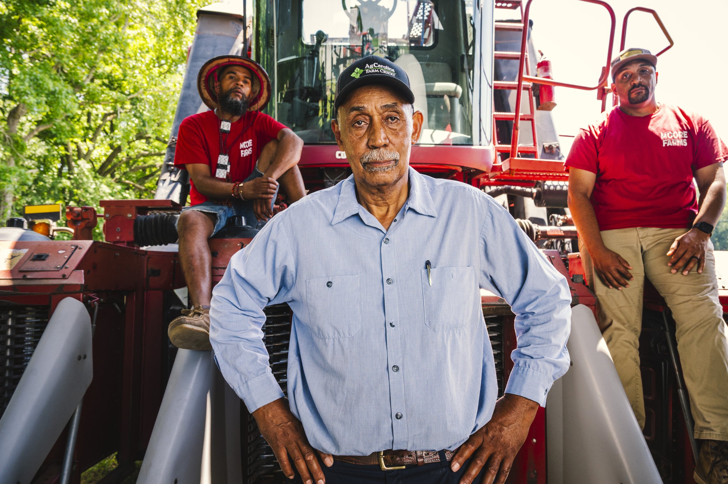 Group of four men standing near a red tractor with slides in a green outdoor setting. The man in the foreground has a mustache, wearing a light blue shirt and a black cap. The other three men are in the background, one wearing a wide-brimmed hat, ano