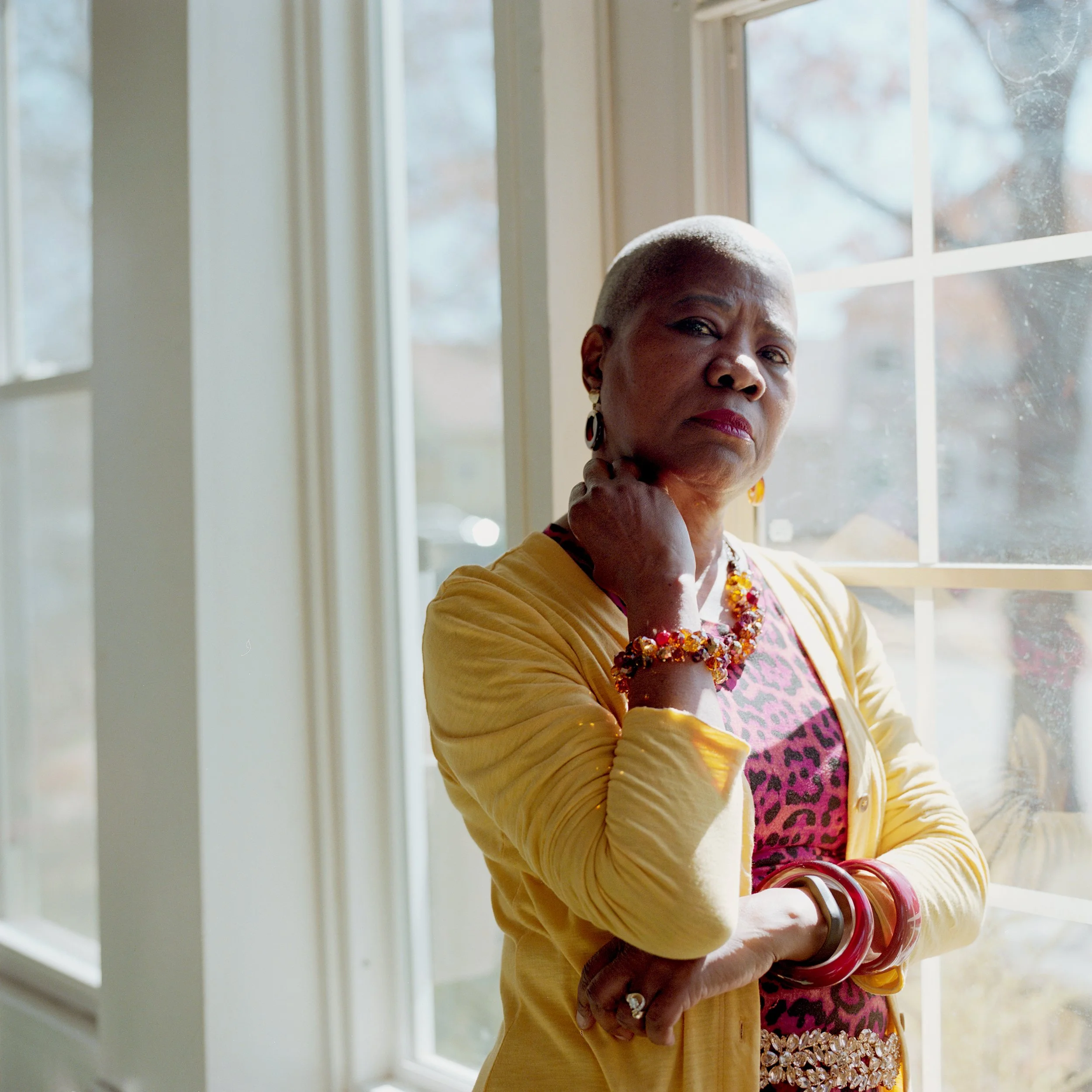 A confident woman with a shaved head standing indoors near a window, wearing colorful jewelry and a patterned top, looking directly at the camera. She is contemplating the loss of her brother to COVID-19.