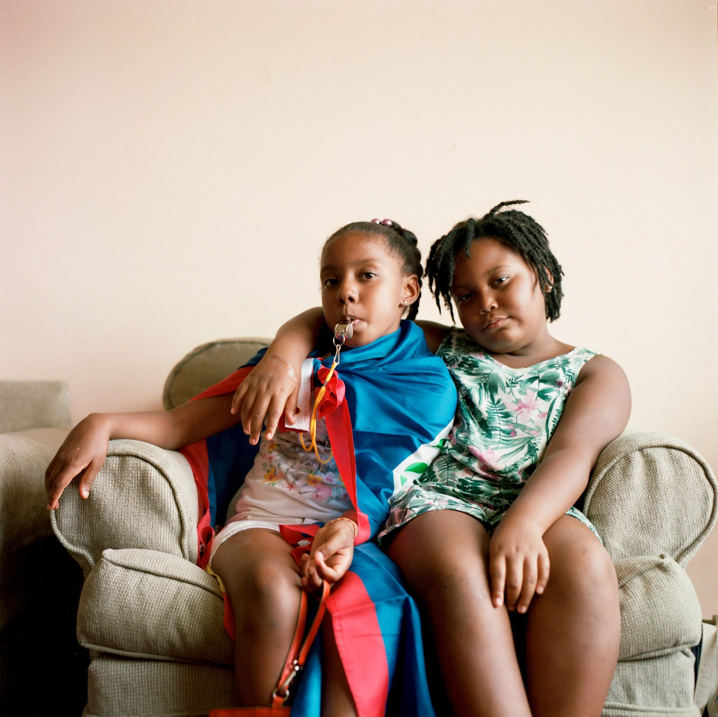 Two young girls sitting together on a beige couch, one with her arm around the other. The girl on the left is wearing a blue and red outfit, with a pacifier in her mouth and a red ribbon tied around her arm. The girl on the right is wearing a green f