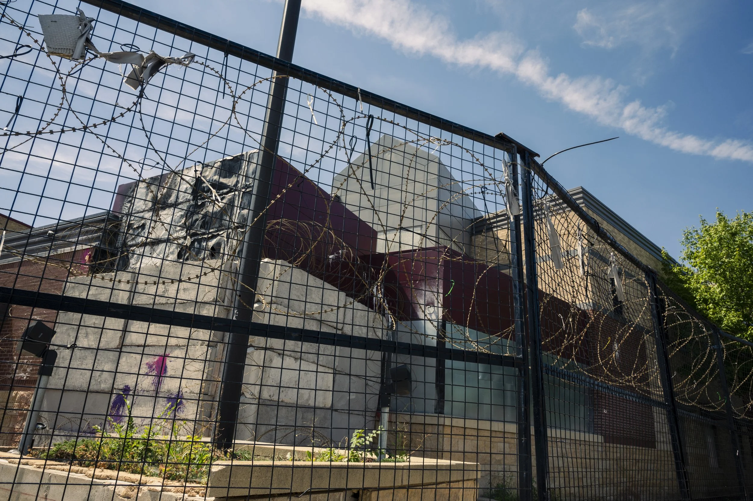 A chain-link fence topped with barbed wire surrounds a graffiti-covered wall with underground storage tanks behind it, against a blue sky with clouds and some trees.
