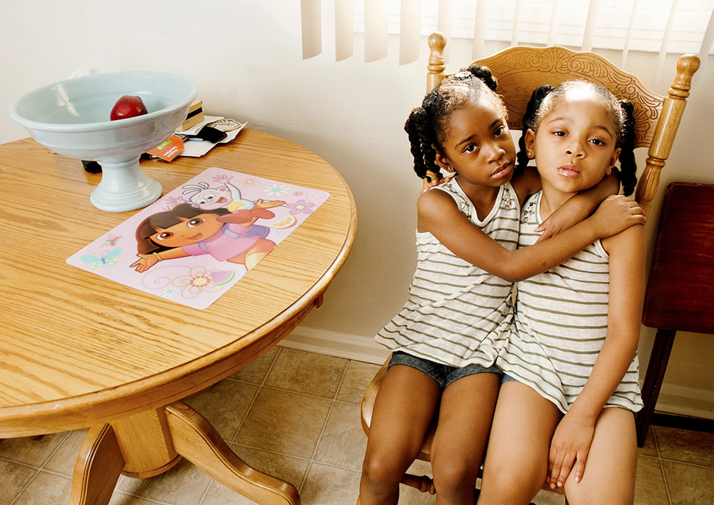 Two young girls sitting close together on a wooden chair, hugging each other, in a kitchen setting with a table nearby that has a bowl with a red apple and some paper items, and a Dora the Explorer themed placemat.