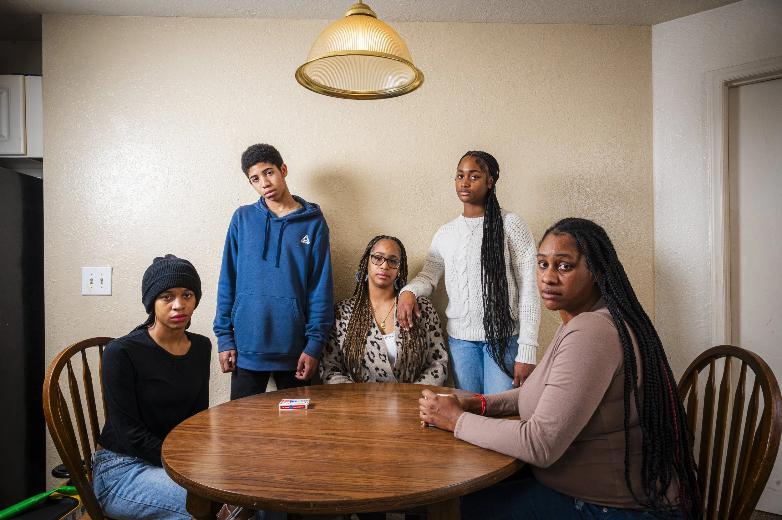 Five people, four women and one man, sitting and standing around a wooden dining table with a pack of playing cards, in a room with beige walls and a hanging lamp.