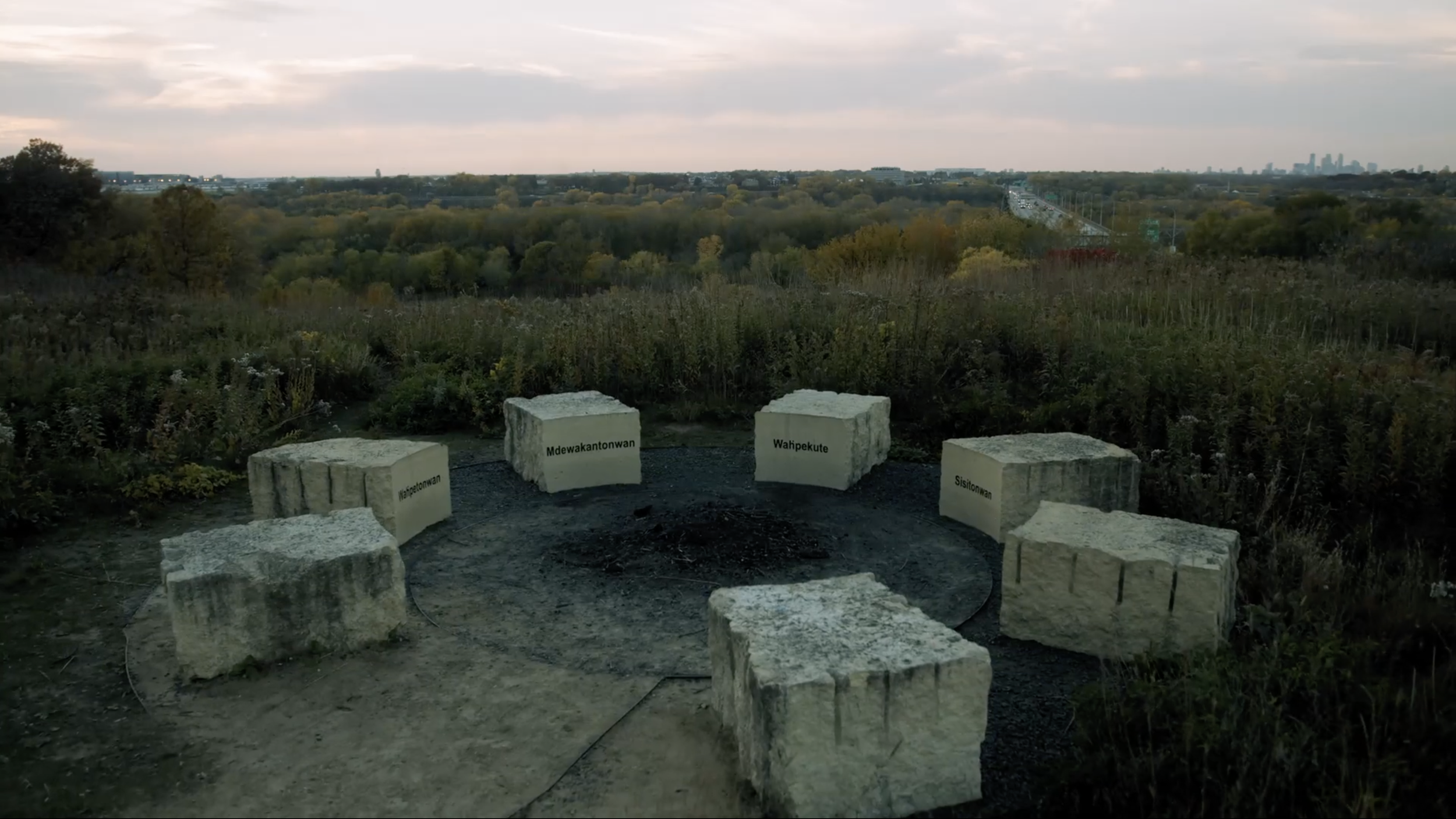 A circular stone seating area with six large rocks, each labeled with different place names, overlooking a vast landscape with a city skyline in the distance at sunset.