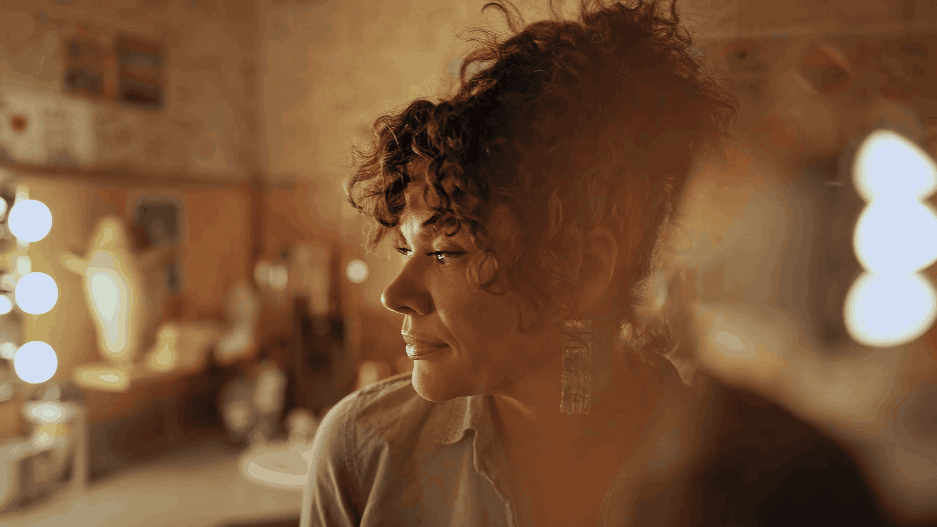 Close-up of a woman with curly hair and earrings in a warmly lit room with blurred background.