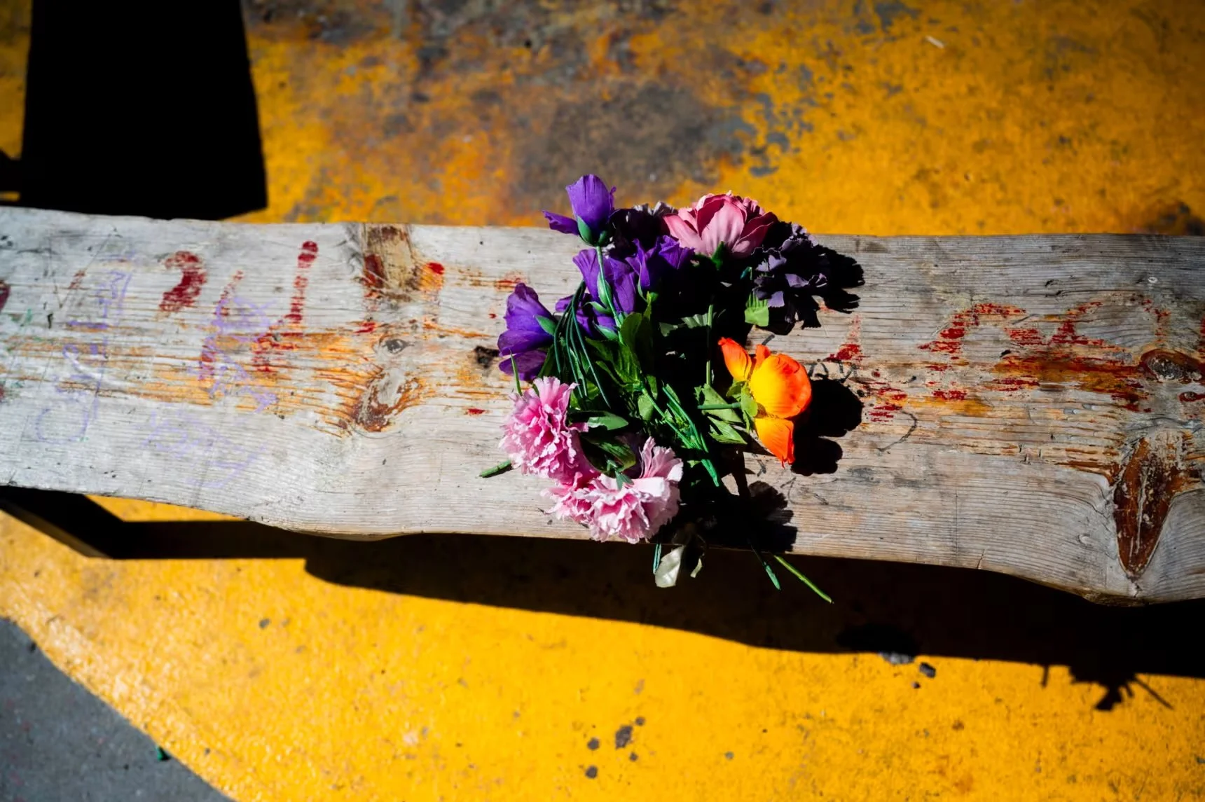 A bouquet of colorful flowers including purple, pink, yellow, and orange blooms placed on a weathered wooden surface on a bright yellow ground.
