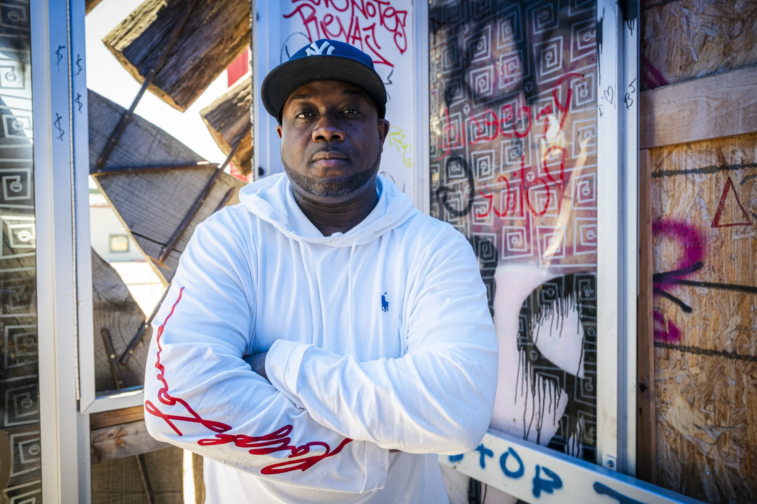A man wearing a black New York Yankees baseball cap and a white hoodie with red and blue writing, sitting with his arms crossed in front of a colorful graffiti-covered wall and wooden boards with graffiti and signs in the background.