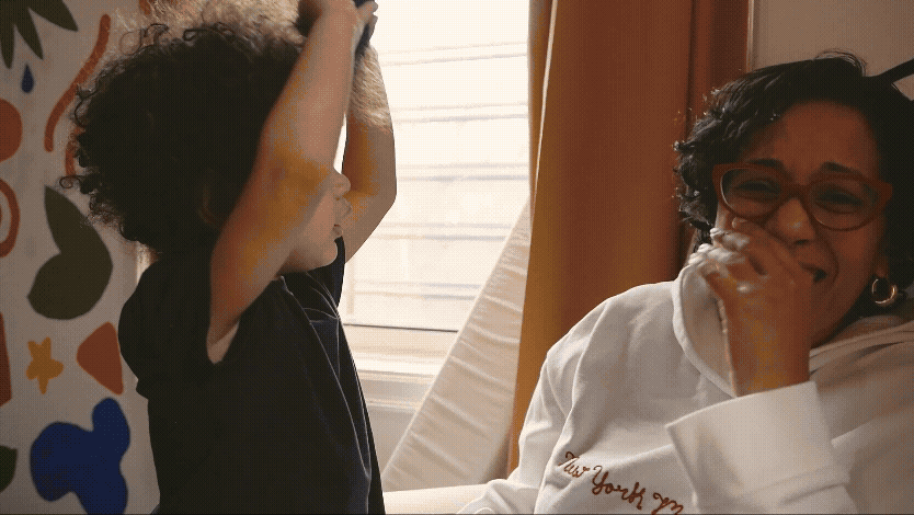 A Latina woman and her young son are sitting near a window with sunlight. The woman is smiling, wearing glasses and a white hoodie, while her son has curly hair, is wearing a black shirt, and is holding her arms up.