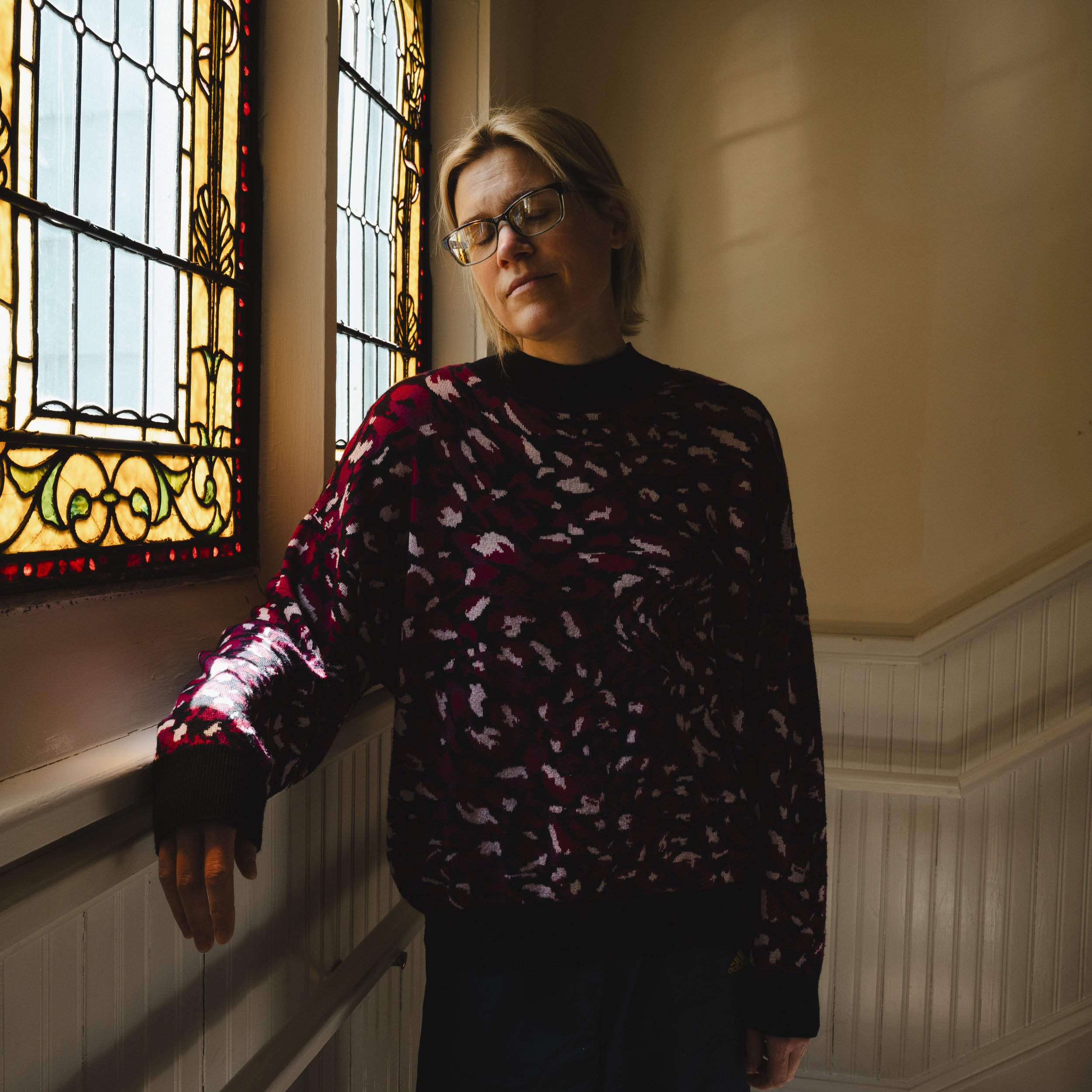 A woman with glasses and short blonde hair, wearing a patterned sweater, standing indoors near a stained glass window with her eyes closed.