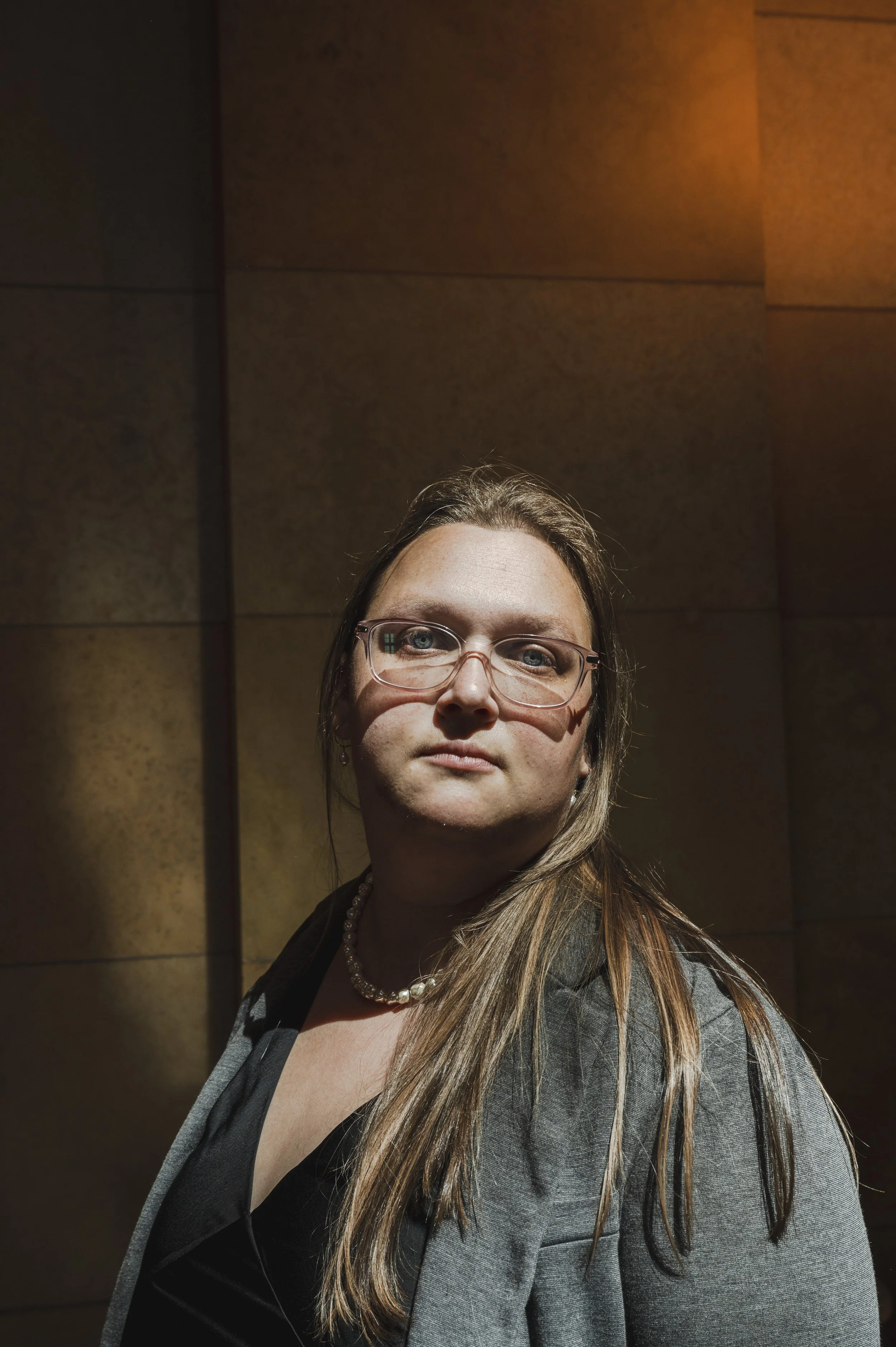 A woman with long, light brown hair, wearing glasses and a pearl necklace, stands in front of a dark, wooden wall.