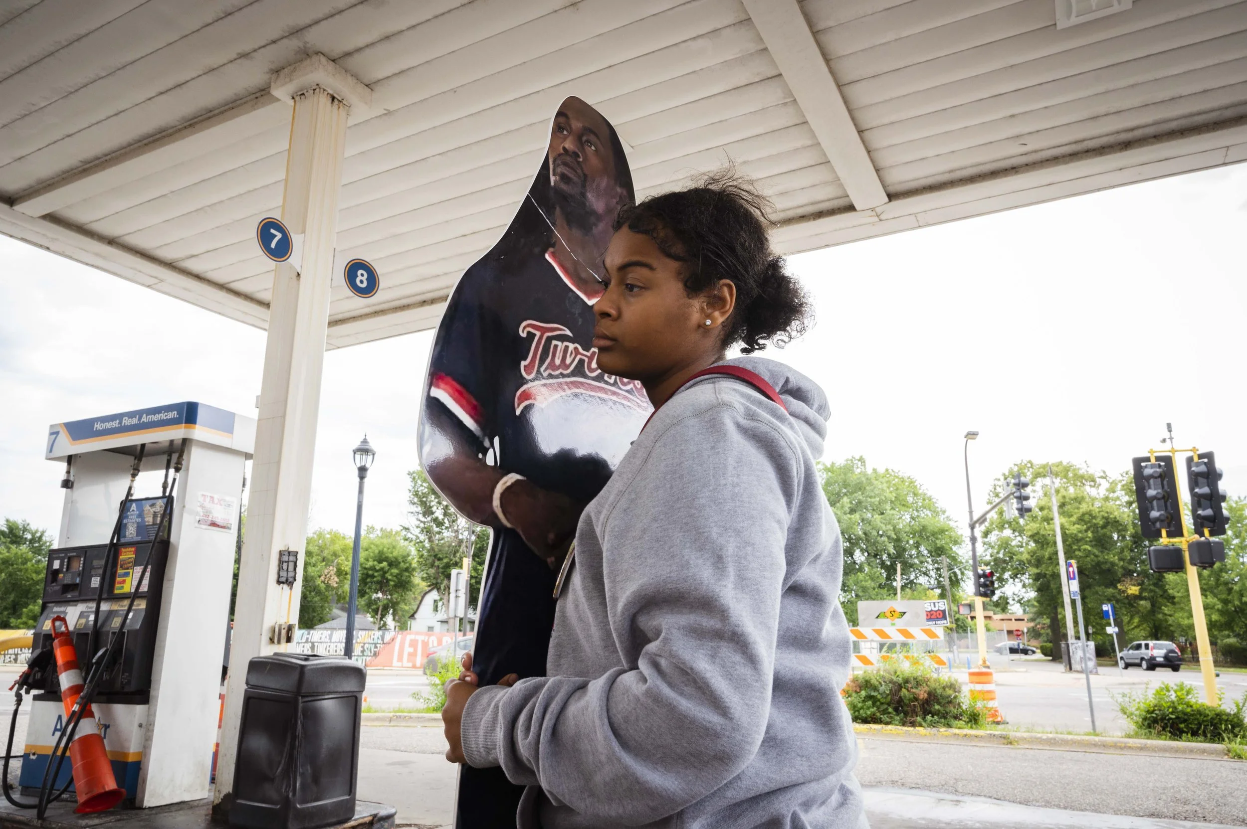 A young woman standing at a gas station, holding a large cardboard cutout of a man, with gas pumps and a parking lot in the background.